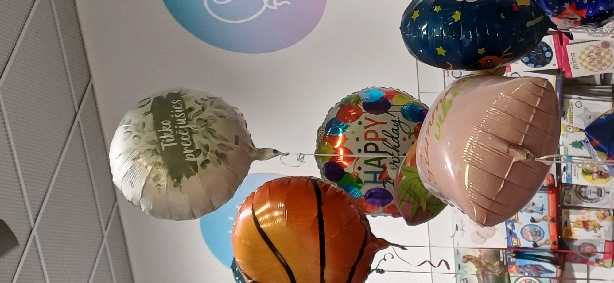 A medium shot captures a cluster of balloons hanging indoors, likely in a party supply store. The balloons are of various shapes and designs. The most prominent is a large, silver, round balloon with a green leafy design and text that appears to read "Tikko prečjūšies." To its left, a balloon designed to resemble a basketball is visible. Below these, a multicolored round balloon displays the text "HAPPY birthday!" A pink, heart-shaped balloon with a textured pattern is positioned in the foreground, partially obscuring a dark blue, star-patterned balloon. In the background, shelves stocked with packaged party supplies and toys are visible, suggesting a retail environment. The lighting is bright and even, typical of an indoor commercial space. The ceiling, constructed of white acoustic tiles, is visible at the top of the frame. The exact location is unknown.