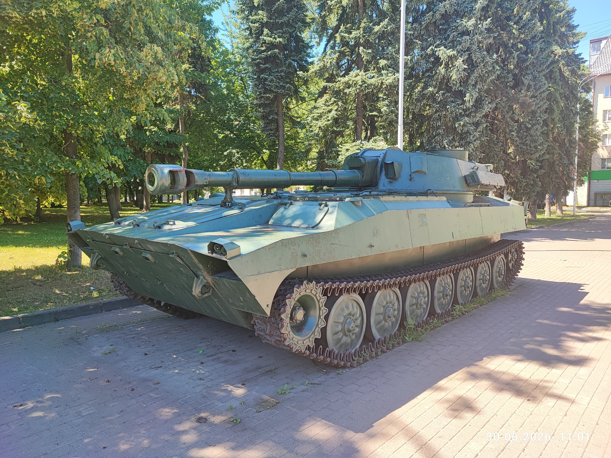This is a full-shot, eye-level, outdoor photograph of a military tank on display on a paved surface. The tank is painted in a camouflage green color. It has a long barrel and a turret. The tank is positioned at an angle, facing slightly to the left of the camera. The background consists of lush green trees, with some buildings visible in the distance on the right. The sky is blue and clear. Sunlight casts shadows on the paved ground. A date and time stamp, "30-06-2026 11:01", is visible in the lower right corner of the image. The location is identified as the city of Lytkarino, Russia. There are no people or animals visible in the image.