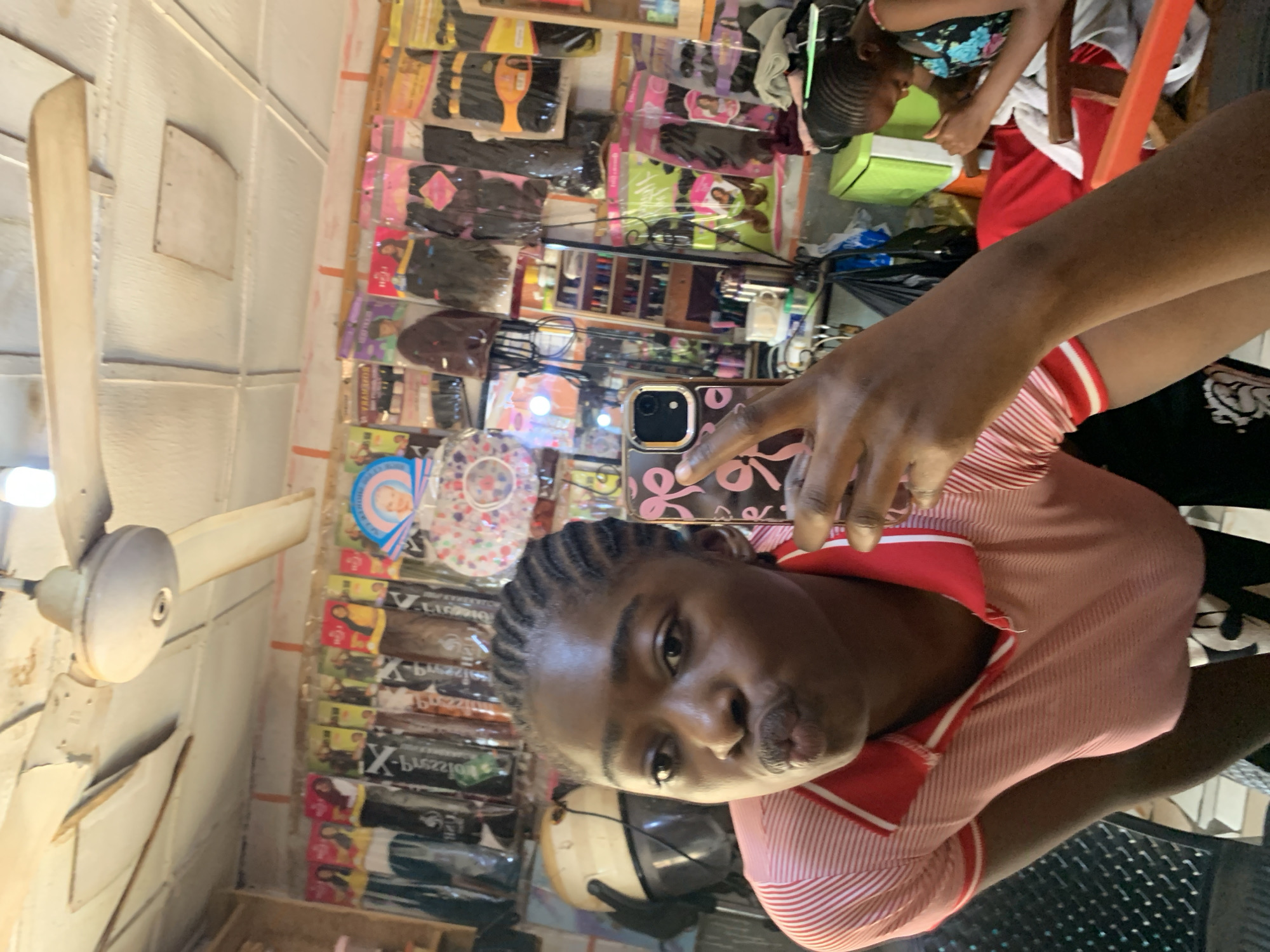 The image shows a young Black woman taking a selfie in a shop. She is pouting and wearing a pink and white striped polo shirt. The shop appears to be a hair salon, with shelves of hair extensions and products visible in the background. There is also a hair dryer and a mirror. Another young Black woman can be seen in the background, working on someone's hair. The setting is likely a small, local business in Rafin Par, Nigeria, given the products displayed and the style of the shop. The overall atmosphere seems informal and busy.