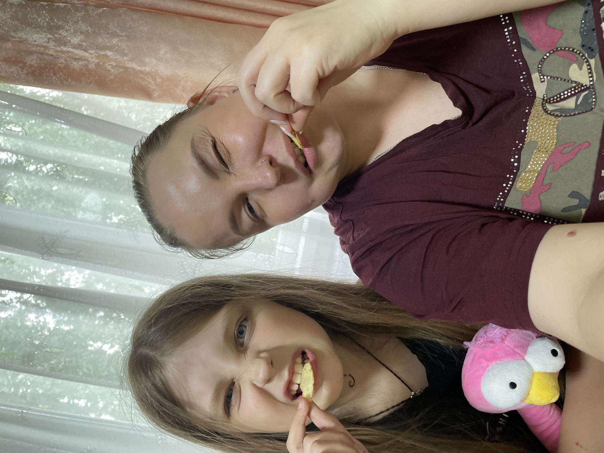 The image captures two individuals, a young girl and a woman, appearing to be in a residential setting. Both are holding and seemingly eating potato chips. The young girl is positioned in the lower portion of the frame, with long hair and a playful expression. Beside her is a pink stuffed animal, possibly a flamingo. The woman is positioned above the girl, also eating a chip. The background includes curtains and what appears to be a window. There is no verifiable evidence within the image to suggest the location is Izhevsk, Russia.