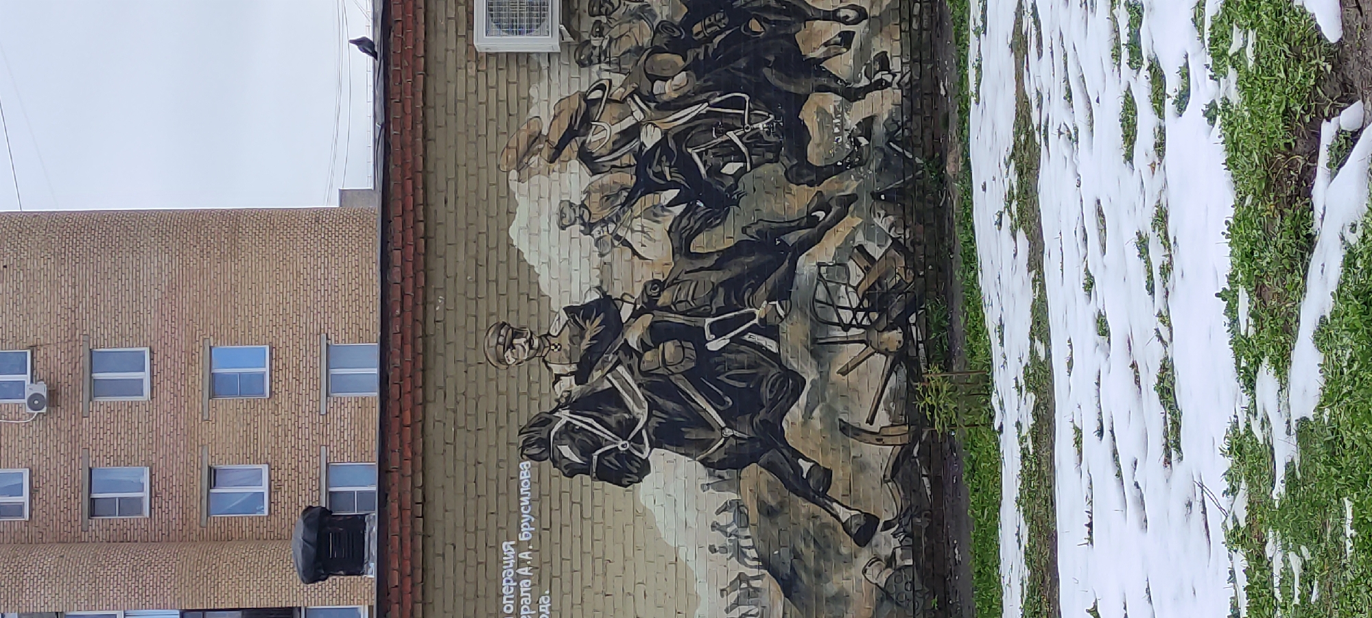 An outdoor image captures a large mural painted on the side of a light brown brick building, depicting a historical military scene. The mural, rendered in shades of black, grey, and brown, portrays a dynamic battle or charge with multiple soldiers on horseback, presumably cavalry, amidst what appears to be a chaotic battlefield. A prominent figure, likely a general or leader, is depicted on a horse in the foreground, wearing a helmet and military attire.
To the left of this central figure, text is visible on the mural, which reads in Cyrillic: "операция генерала А.А. Брусилова" (operation of General A.A. Brusilov), indicating that the artwork commemorates the Brusilov Offensive, a major World War I military operation.
The building on which the mural is painted is made of light brown brick and features numerous rectangular windows, some with air conditioning units visible on the facade. Above the mural, the sky is overcast, suggesting daytime.
To the right of the mural, a strip of ground is visible, covered in patches of white snow alternating with vibrant green grass, implying a winter or early spring setting. The overall scene is set in an urban or suburban environment in Fryazino, Russia.