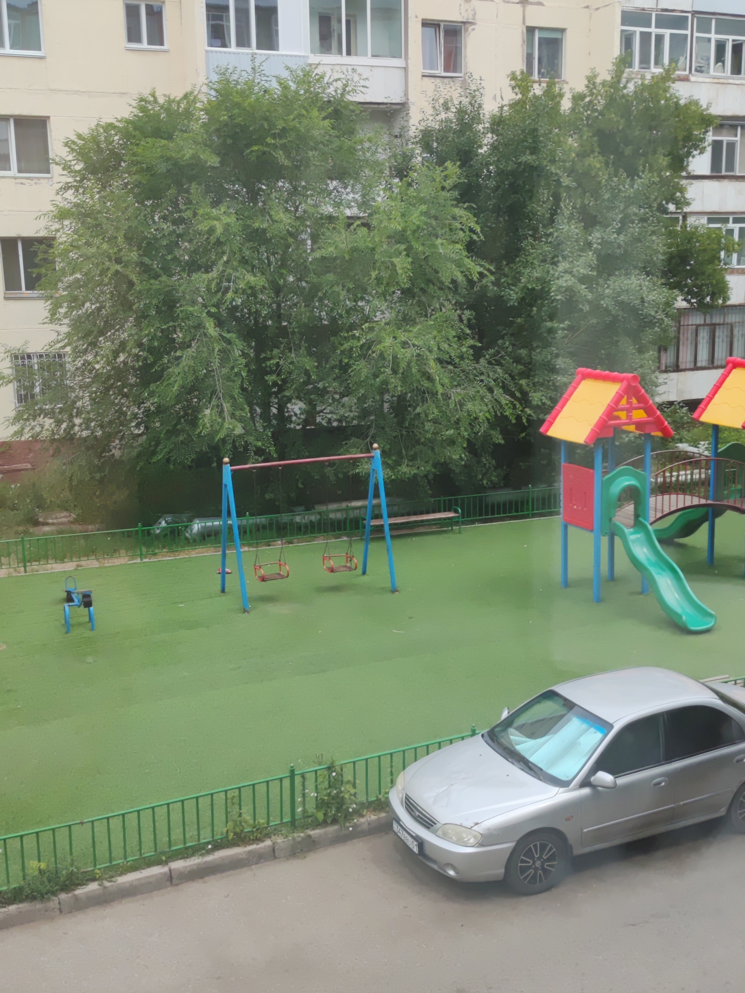 The image shows a playground and a street in Astana, Kazakhstan. The playground has green turf, a swing set with two red swings, a small blue seesaw, and a colorful play structure with a green slide. Adjacent to the playground is a silver sedan parked on an asphalt street. Trees and a multi-story building are visible in the background. The scene appears to be set during the daytime, with overcast or cloudy conditions suggested by the lighting.