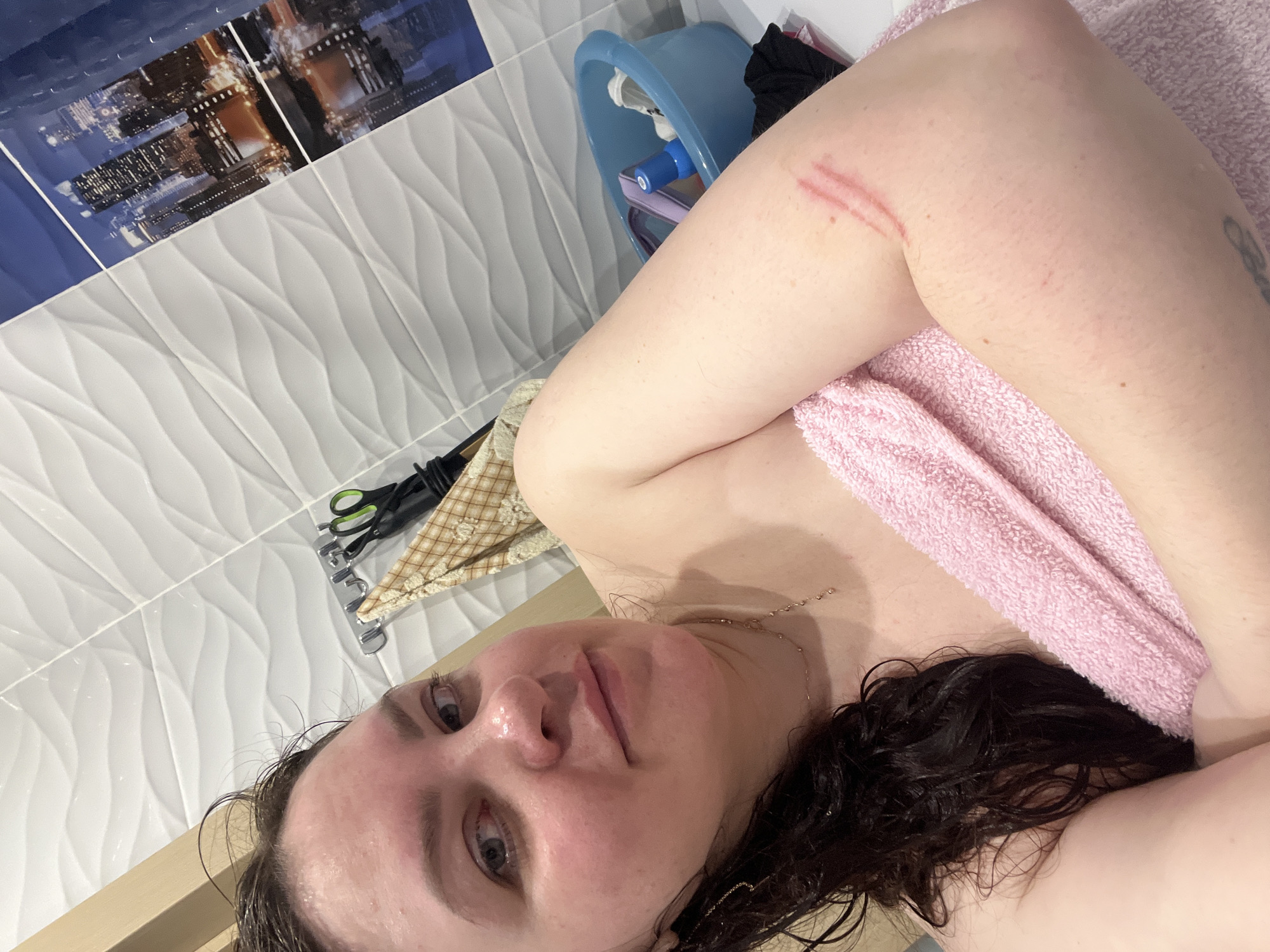 A slightly rotated, medium close-up selfie captures a person, likely female, with wet, dark brown hair, looking directly at the camera with a neutral expression. She is bare-shouldered, with a light pink towel draped across her chest and right shoulder. A thin gold chain necklace hangs around her neck.
Notable red marks or scratches are visible on her upper right arm, appearing as two distinct parallel lines and a fainter third line. Her skin shows a slight flush, possibly from a recent shower or bath.
The background indicates an indoor setting, likely a bathroom in Perm, Russia. The wall features two distinct tile patterns. The upper section displays dark blue, smooth tiles with a printed cityscape motif showing illuminated buildings at night. Below this, the majority of the visible wall is covered with white, wavy-textured tiles arranged horizontally.
To the upper right, behind her arm, a light blue plastic bucket containing some items is partially visible. Further to the left and lower, a pair of black-handled scissors and a piece of checkered fabric are seen hanging on a metallic rack against the white tiles. The lighting in the scene is consistent and even, suggesting indoor artificial illumination. No legible text is visible in the image.