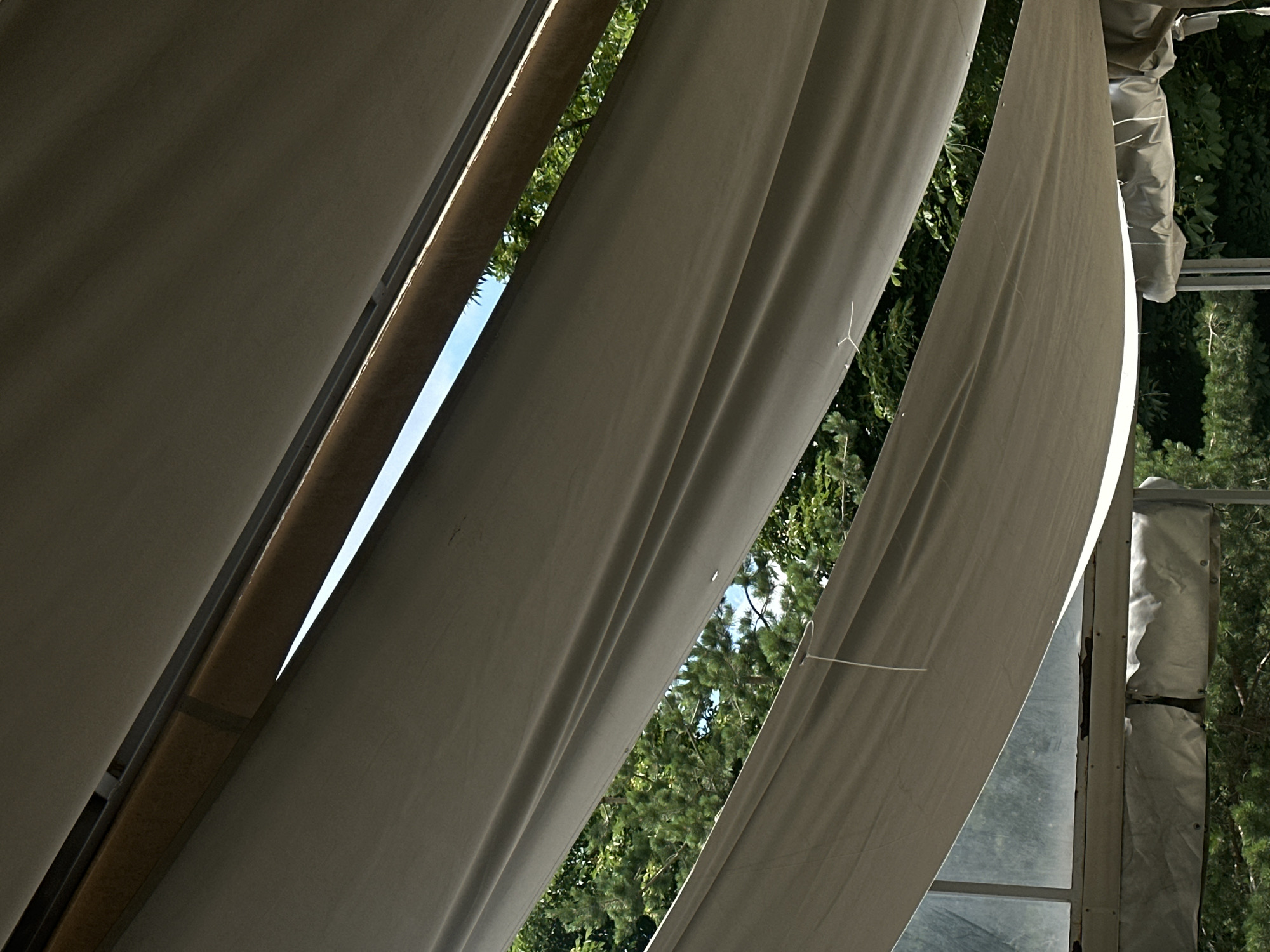 The image shows a view looking upwards through the gap of a tent or canopy structure. Multiple layers of light-colored fabric are draped in a way that creates shadows and highlights. Through a significant gap between the fabric layers, green trees and a bright blue sky are visible, suggesting an outdoor setting. The structure appears to be made of fabric supported by metal poles, with some visible hardware and ties. The overall impression is one of an outdoor shelter, possibly a temporary structure. The location is not identifiable from the image content.