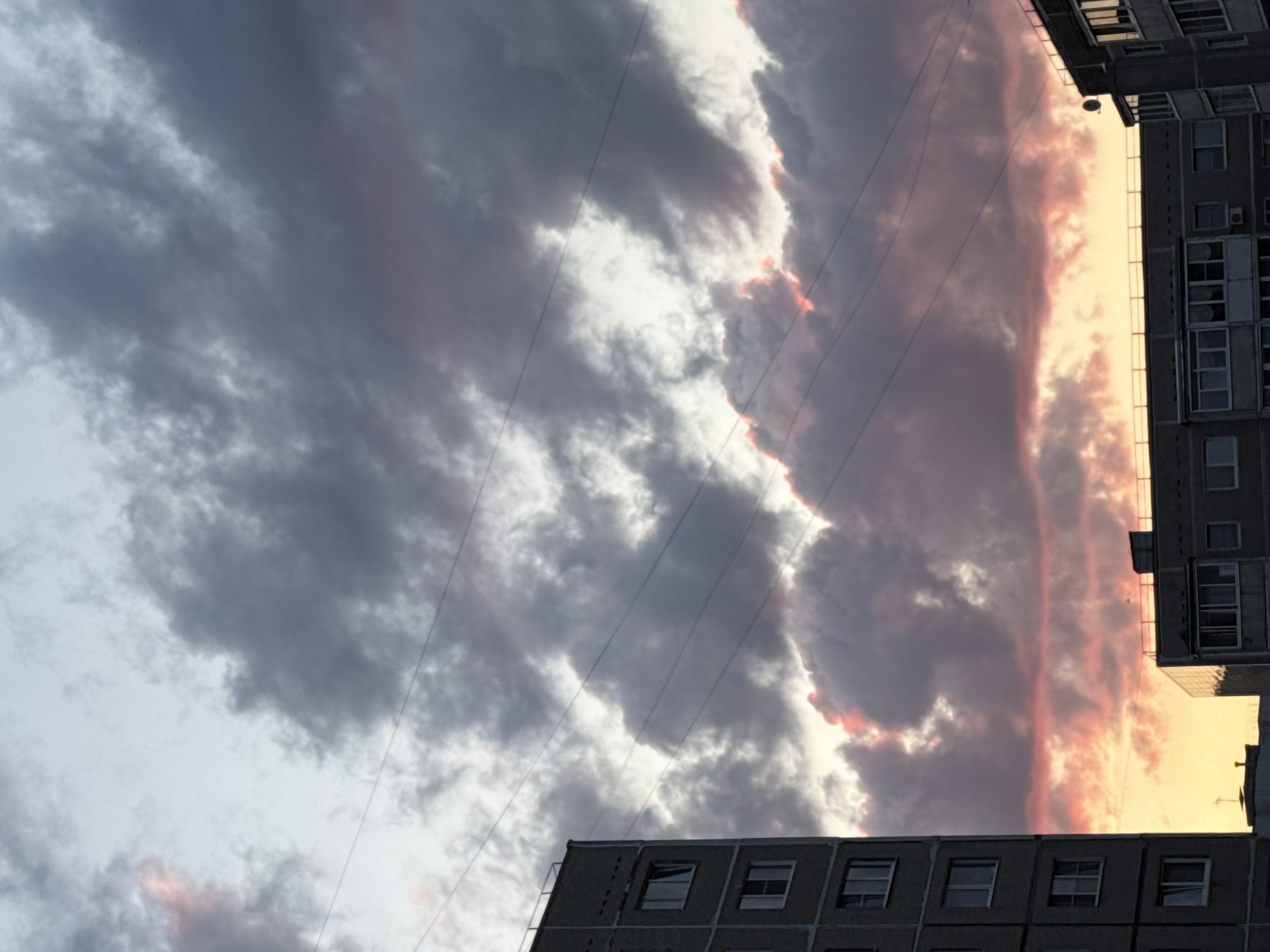 The image, oriented vertically, captures a cityscape in Chaikovsky, Russia. Two gray concrete buildings frame the sky. One building is situated at the bottom center, revealing several rows of windows. The second building is visible on the upper right, appearing taller with multiple floors and windows. Above these buildings, the sky is filled with a dynamic cloud formation. The clouds are a mix of gray and white, with a strip of pinkish-orange light peeking through, likely indicating either sunrise or sunset. Several power lines cut through the sky, adding a linear element to the composition. The lighting suggests that the image was taken during twilight hours. No people or specific activities are visible.