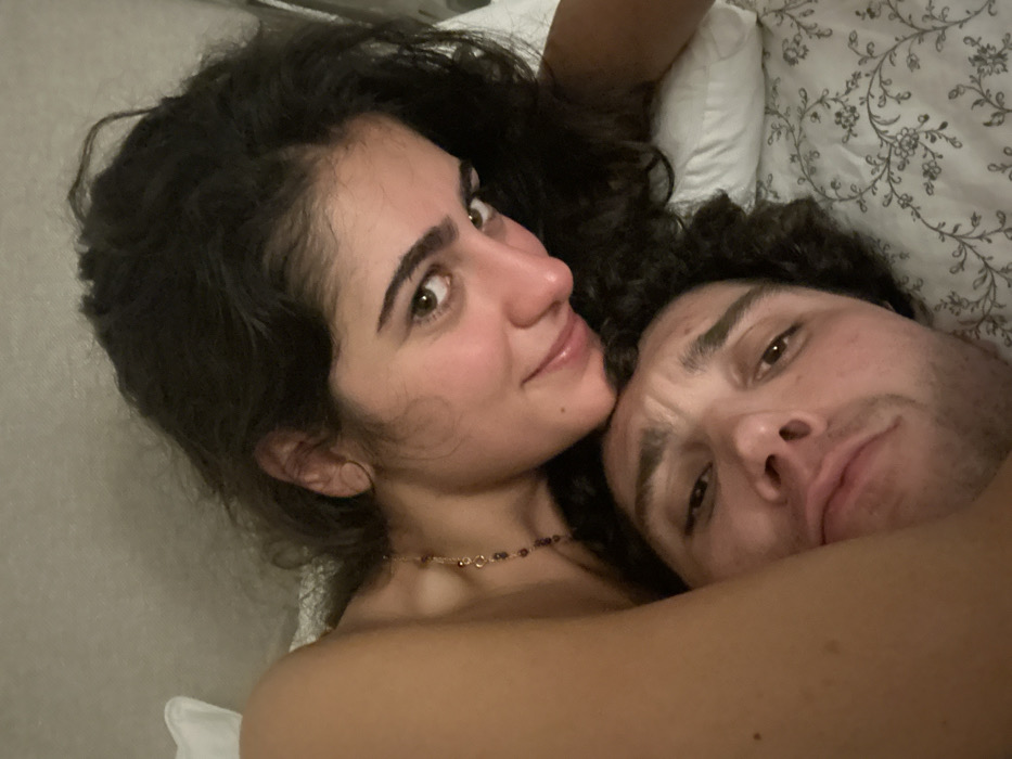 A cozy, close-up snapshot shows a woman and a man lying in bed. The woman, with dark curly hair and a gentle smile, gazes directly at the camera. She is wearing a delicate beaded necklace. Next to her, the man is nestled closer, his face bearing a slightly weary expression. The bedding includes a white pillow with a subtle floral pattern, providing a soft background. The lighting is somewhat dim.