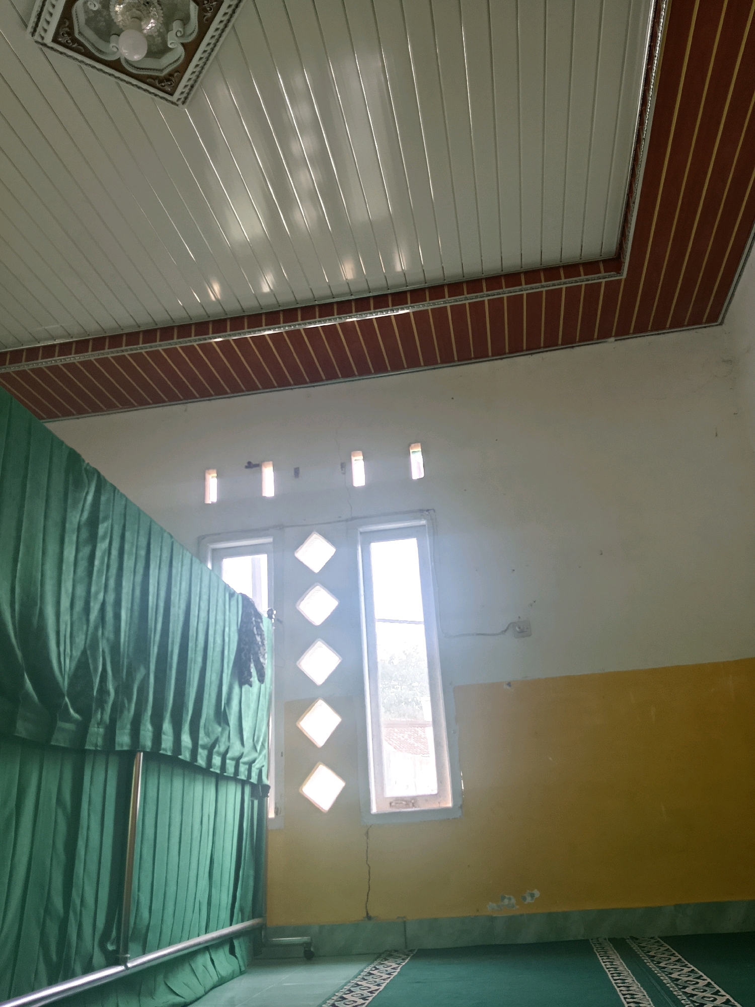 The image depicts the interior of a room, likely in Negeri Agung, Indonesia. The ceiling is white and paneled, with a decorative brown and gold border. A light fixture is visible on the ceiling. The walls are a pale cream color, with a yellow band along the lower section.
In the left half of the image, a large green curtain is draped, obscuring a significant portion of the room. The curtain appears to be made of fabric and is pleated, with a silver-colored railing in front of it.
To the right of the curtain, a large window or glass door is visible, divided into sections. Some sections are diamond-shaped and contain opaque white inserts. Through the upper portion of the window, trees and buildings can be seen outside.
Above the window, several small rectangular openings are visible in the wall, likely vents or small windows, with light emanating from them.
The floor is covered with a green carpet that has a patterned border.
There are no people visible in the image. The scene appears to be a static interior, possibly a prayer room or a multipurpose room. The lighting suggests it is daytime.