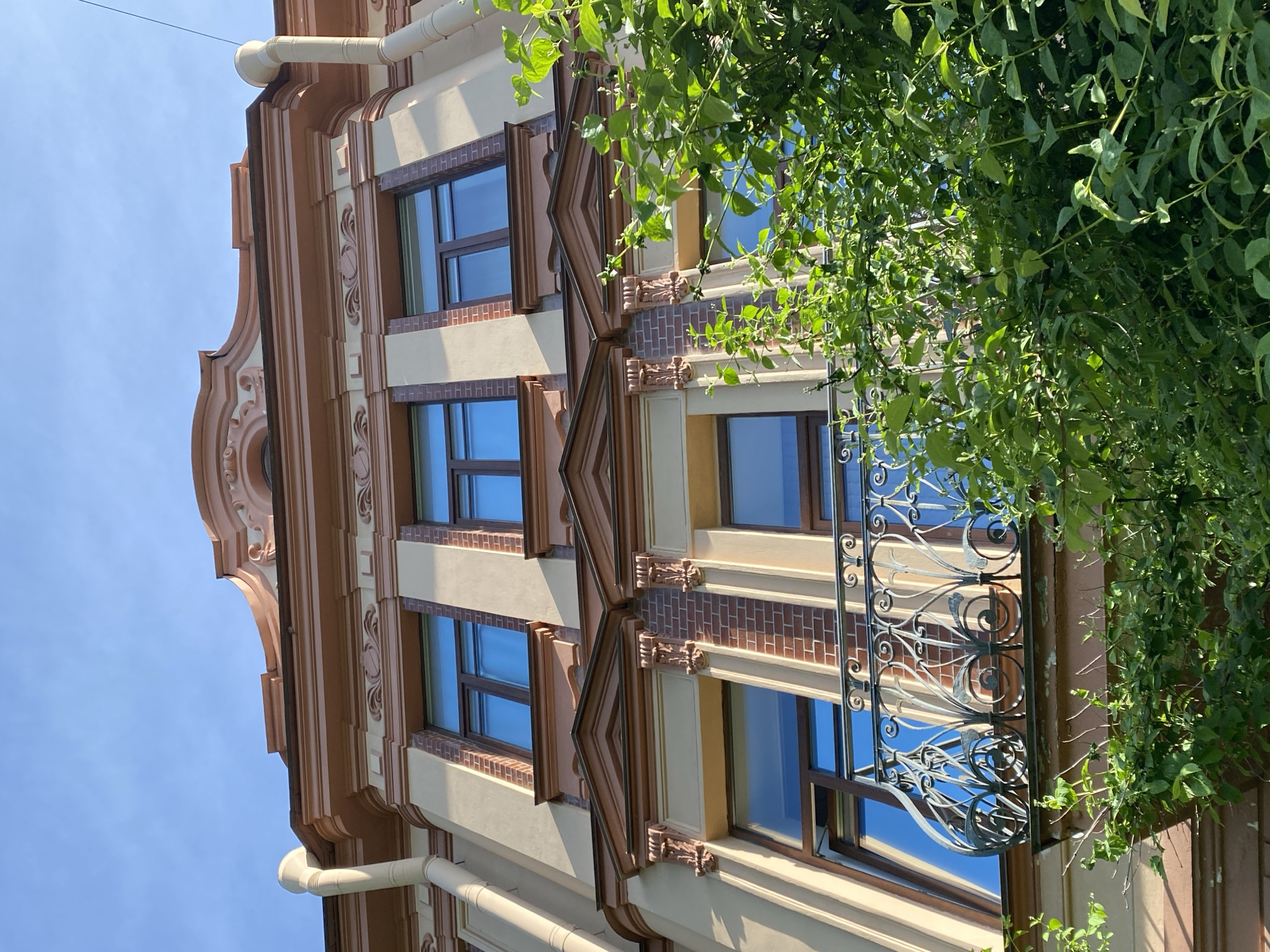 The image captures the exterior facade of a building in Saint Petersburg, Russia, viewed from a low angle. The architectural style incorporates ornate details, with a color palette of beige, brown, and terracotta. The building has multiple windows with dark frames, reflecting the blue sky. A decorative balcony with wrought iron details is partially obscured by green foliage. A downspout runs vertically along the building's edge. The sky is visible in the upper left corner.