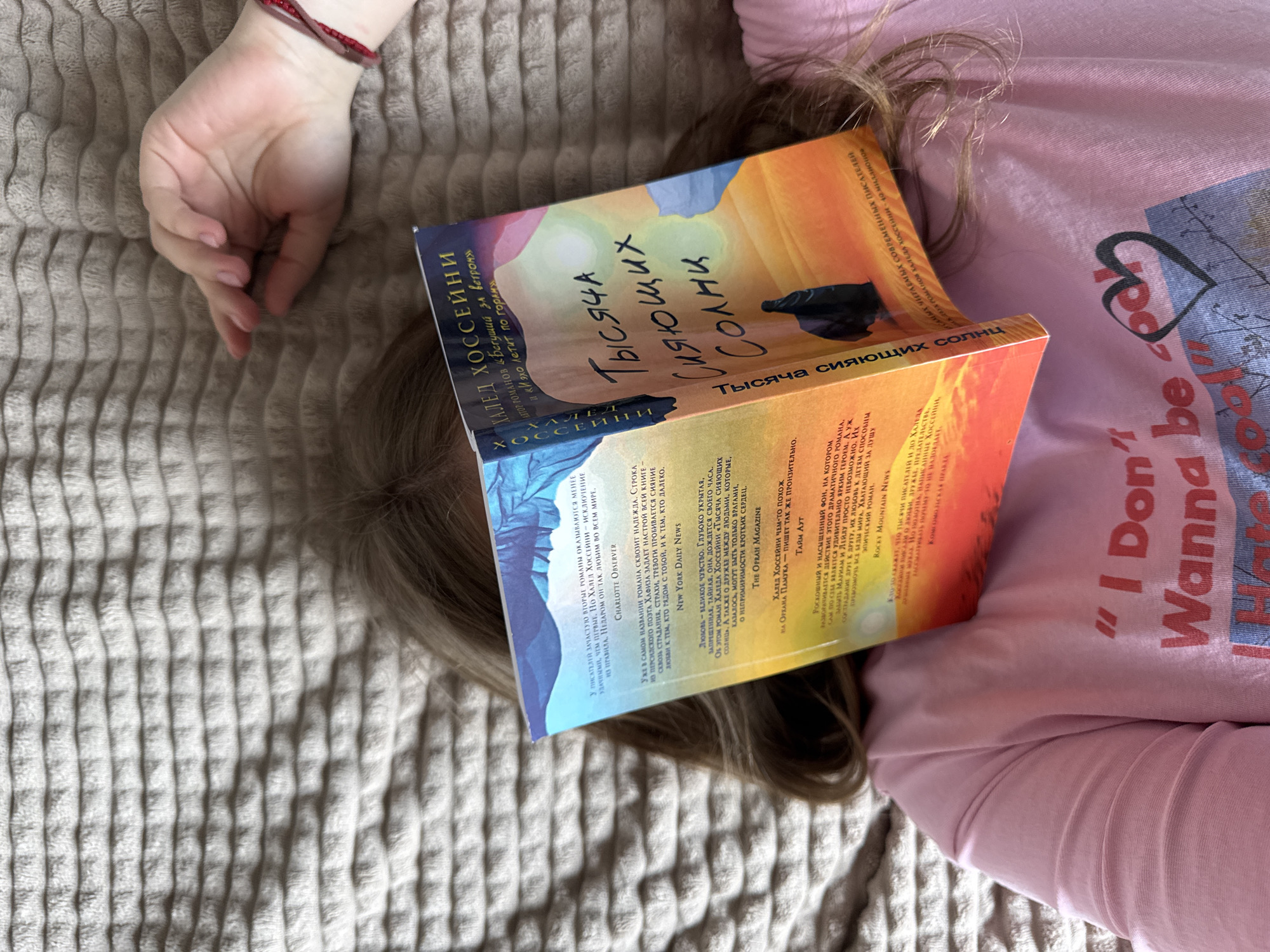 An overhead shot depicts an individual lying supine on a light-colored, horizontally ridged blanket or comforter in Gornyi (Dvurechenskoe S/P), Russia. The person's face is obscured by a partially open book, "Тысяча Сияющих Солнц" (A Thousand Splendid Suns) by Khaled Hosseini, written in Russian. The book's front cover displays a desert landscape under a warm-toned sky with a solitary dark figure. Light brown hair is splayed around the book. A right hand, adorned with a red braided bracelet, rests above the individual's head. The person is wearing a pink short-sleeved top, partially revealing the text "I Don't Wanna be cooL" alongside a heart graphic.