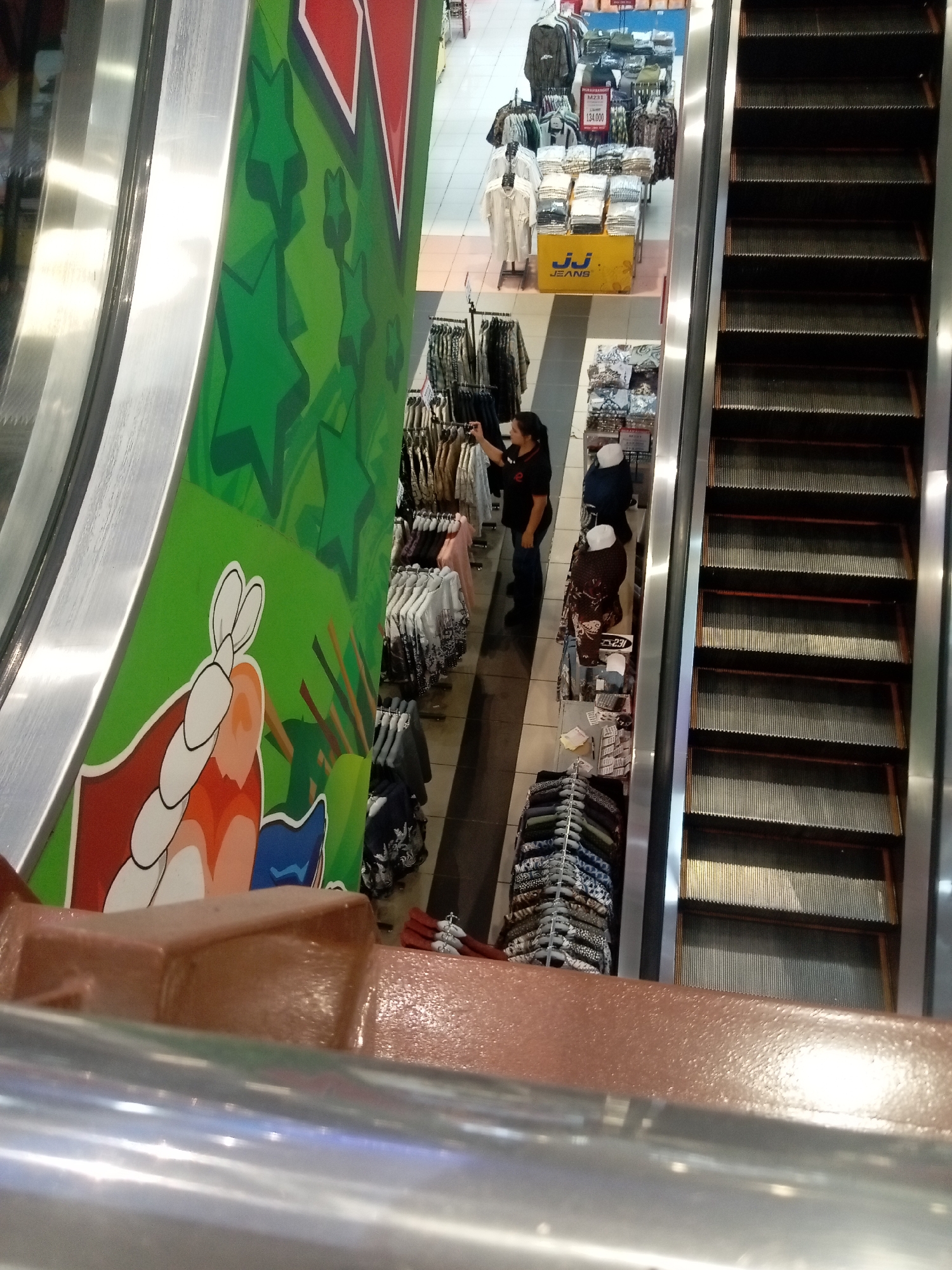An overhead view of an escalator in a department store in Batam, Indonesia. The escalator has dark gray treads and brown side panels. To the left of the escalator is a large, green wall mural decorated with cartoonish depictions of stars and what appear to be shrimps. A section of the mural features a stylized red and white triangular design at the top. In front of the mural and to the left of the escalator, a brown railing with a metallic finish is visible in the foreground.
Below the escalator, the department store floor is visible, displaying various clothing racks and displays. A woman in a black shirt and blue jeans is standing near a rack of clothes, holding a camera and appearing to take a photo. There are several mannequins dressed in traditional Indonesian batik clothing. One display features folded shirts with a yellow sign that says "JJ JEANS". Another section shows stacks of folded clothing on shelves. Red and white flags or banners are visible hanging in the background. The store appears to be well-lit, with fluorescent lighting overhead.
