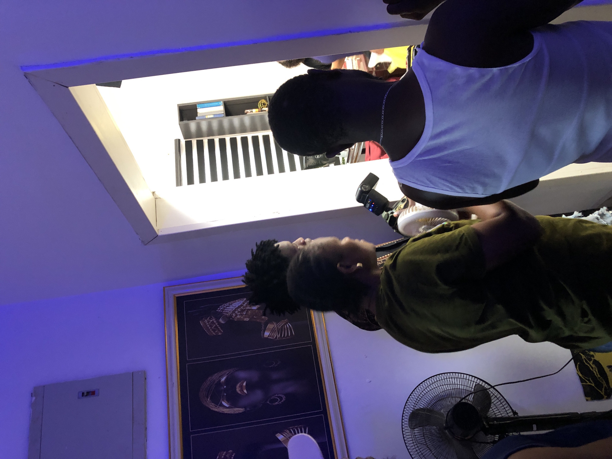 The image captures an indoor scene with two individuals and distinctive modern decor, viewed from a high angle looking down. In the foreground, on the right, a person with short dark hair and wearing a white tank top is visible from the back. They have a silver chain around their neck and are holding a camera with an attached external flash unit, suggesting they are engaged in photography or videography. Below and to their left, another person wearing a dark green shirt is partially visible, also looking in the general direction of the scene.
The room features a large framed artwork on the left wall, displaying multiple stylized portraits or busts with elaborate, gold-toned headpieces against a dark background. To the left of this artwork, a white rectangular panel, possibly an electrical box, is mounted on the wall. A ceiling fan with a dark grill and blades rests on the floor in the lower right portion of the image.
The space is illuminated by artificial blue light from a strip light along the ceiling, creating a cool ambient atmosphere. An opening in the ceiling in the upper left provides a glimpse into an upper level or adjacent room, revealing a bookshelf stocked with books and a structure with vertical black and white stripes, possibly a decorative screen or railing. The lighting suggests an evening or night setting. No legible text is visible.