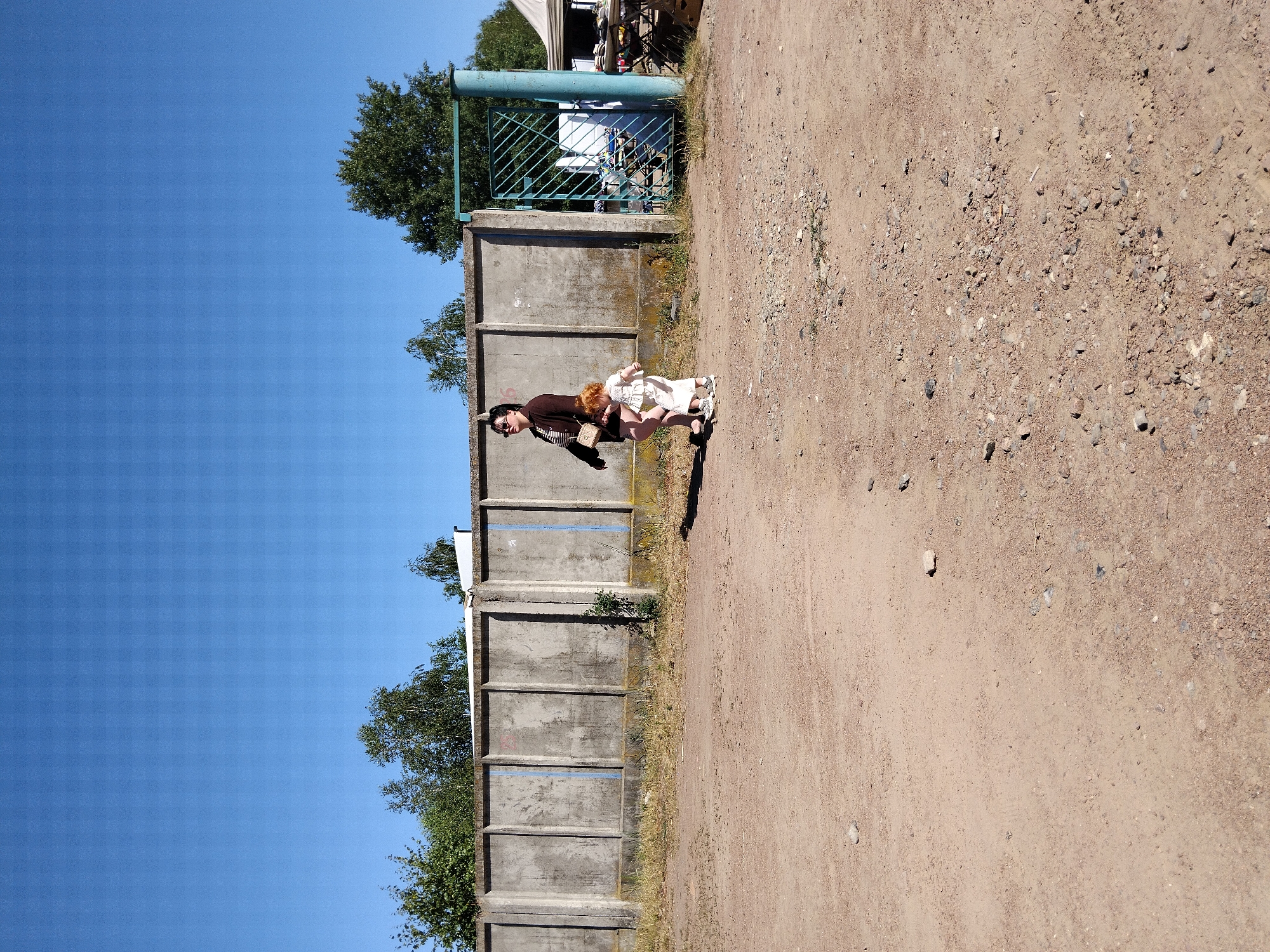A woman and a young child stand on a dirt path in front of a tall, grey concrete wall with vertical divisions. The woman, on the left, wears a dark brown jacket over a striped shirt and dark shorts, with sunglasses and a light-colored crossbody bag. The child, to her right, has red hair and wears a white outfit. To the right of the wall, a green metal gate is visible. Beyond the gate, a table with items for sale is set up under a white canopy. Trees line the top of the wall and extend beyond it. The ground is covered in dirt and small stones, with sparse, dry grass growing along the base of the wall. The sky is clear and blue. The scene appears to be an outdoor market or storage area in Irshansk, Ukraine.