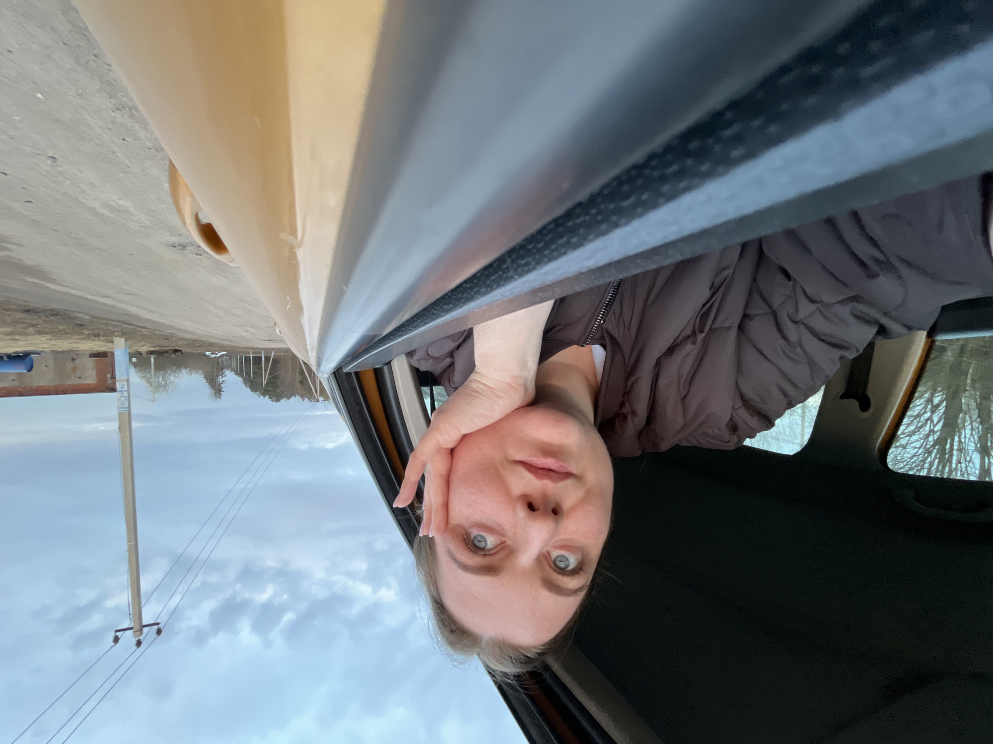 A woman with fair skin and blue eyes is sitting in a car, looking directly at the camera. She has her right hand resting on her cheek. She is wearing a brown quilted jacket. The car's window is down. Outside, utility poles and power lines are visible against a cloudy sky. The location is identified as Pliuskovo, Russia, based on external information.