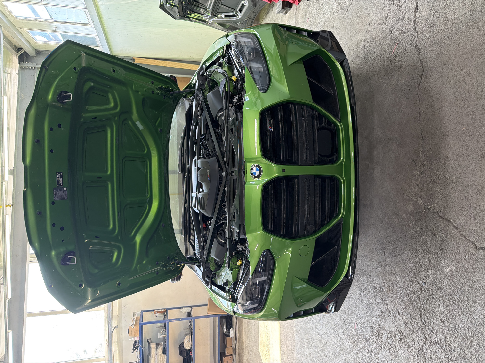 The image shows a green BMW M4 with its hood open, revealing the engine compartment. The car is inside a garage, likely undergoing maintenance. The location is possibly in Burgas, Bulgaria, based on contextual knowledge. The garage appears to be well-lit with fluorescent lighting and has a concrete floor. Shelving units with various objects are visible in the background. The car's hood is fully open, showcasing the engine. The BMW logo is prominent on the front grille. The car's exterior is a bright, metallic green. There are no people visible in the immediate frame.