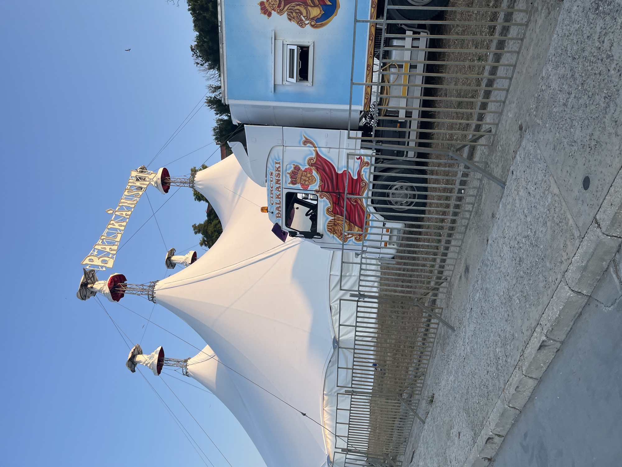 This is a photograph of a circus tent and truck. The tent is white and conical, supported by metal poles. A truck is parked next to the tent, with "CIRCUS BALKANSKI" written on its side in stylized lettering, along with a graphic of a person in regal attire. The truck also has a blue trailer attached. The scene appears to be outdoors, with a clear blue sky and some trees in the background. A metal fence runs along the right side of the image. The photograph is taken at an angle, suggesting the viewer is looking up at the tent and truck. The lighting suggests it is daytime, likely late afternoon or early morning due to the quality of light. There is no visible activity taking place, and the scene appears to be set up, possibly before or after a performance. The location is identified as Nesebar, Bulgaria.