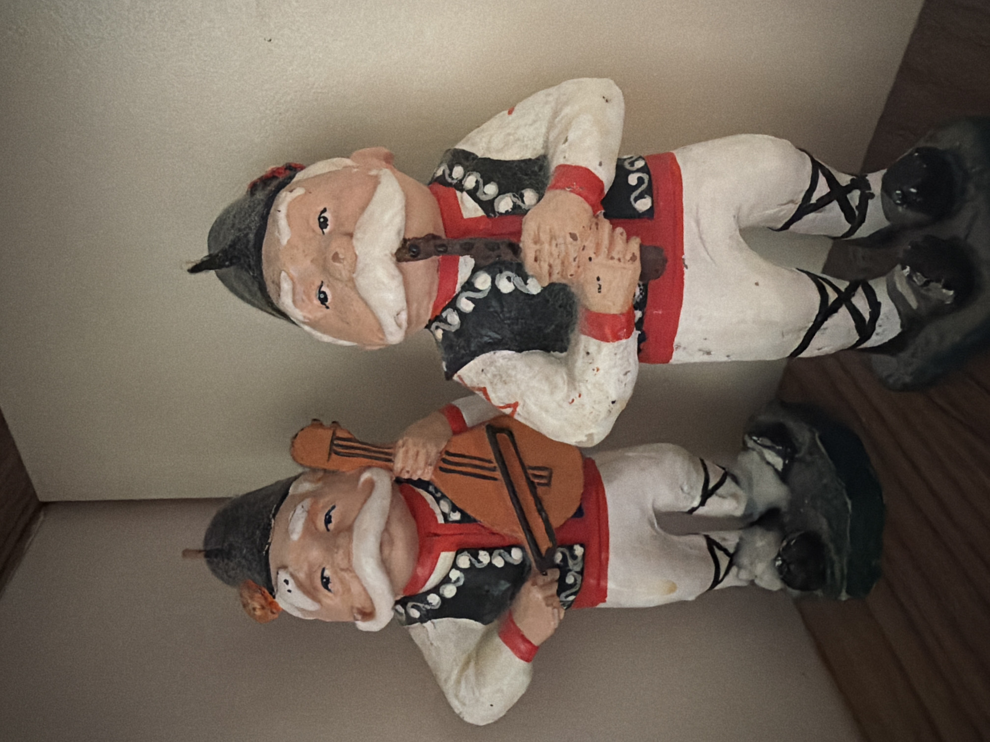 Two static, painted figurines of men in traditional Bulgarian attire stand on a wooden surface, against a plain wall. The figurine on the left, positioned slightly forward, is playing a mandolin-like instrument with a bow. The figurine on the right holds a flute-like instrument to its mouth. Both figures wear hats, white shirts with red collars and vests, white pants with black criss-cross designs on the lower legs, and dark shoes. The background appears to be an interior corner. The specific location in Pazardzhik, Bulgaria cannot be determined from the provided image.