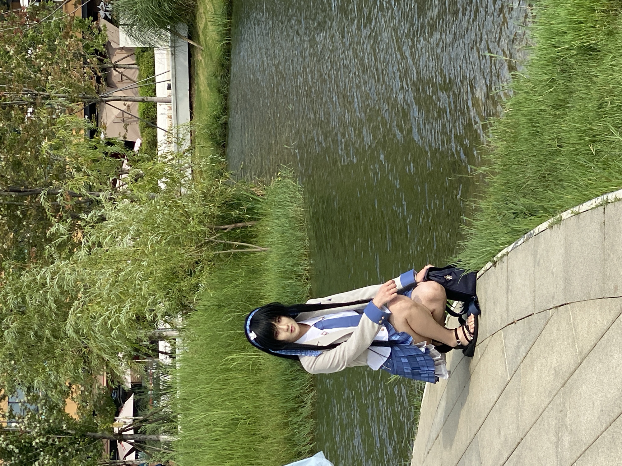 A person dressed in a school uniform is crouching on a paved pathway next to a body of water. The person has black hair with a blue ribbon and white flowers in it. They are wearing a light-colored blazer over a white shirt and blue tie, a blue plaid skirt, and black sandals. A dark backpack is on the ground beside them. The pathway curves around the edge of the water, which is surrounded by lush green grass and trees. In the background, there are more trees and some structures that appear to be part of a modern cityscape. The scene is likely located in a park or a landscaped area within Jinniu District, China, given the context provided. The lighting suggests it is daytime, with sunlight casting reflections on the water.