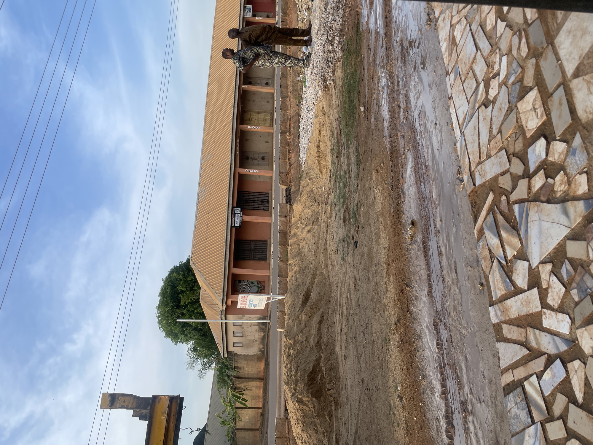 The image depicts a street scene in Narayi, Nigeria. A two-story building with a brown corrugated metal roof stands along the roadside. Two men are standing adjacent to the building and a pile of gravel. The road, with a wet, muddy surface, is adjacent to a sidewalk adorned with irregular tiles. A small sign is planted near the building. Overhead power lines are visible against a partly cloudy sky. A green tree is also near the building.