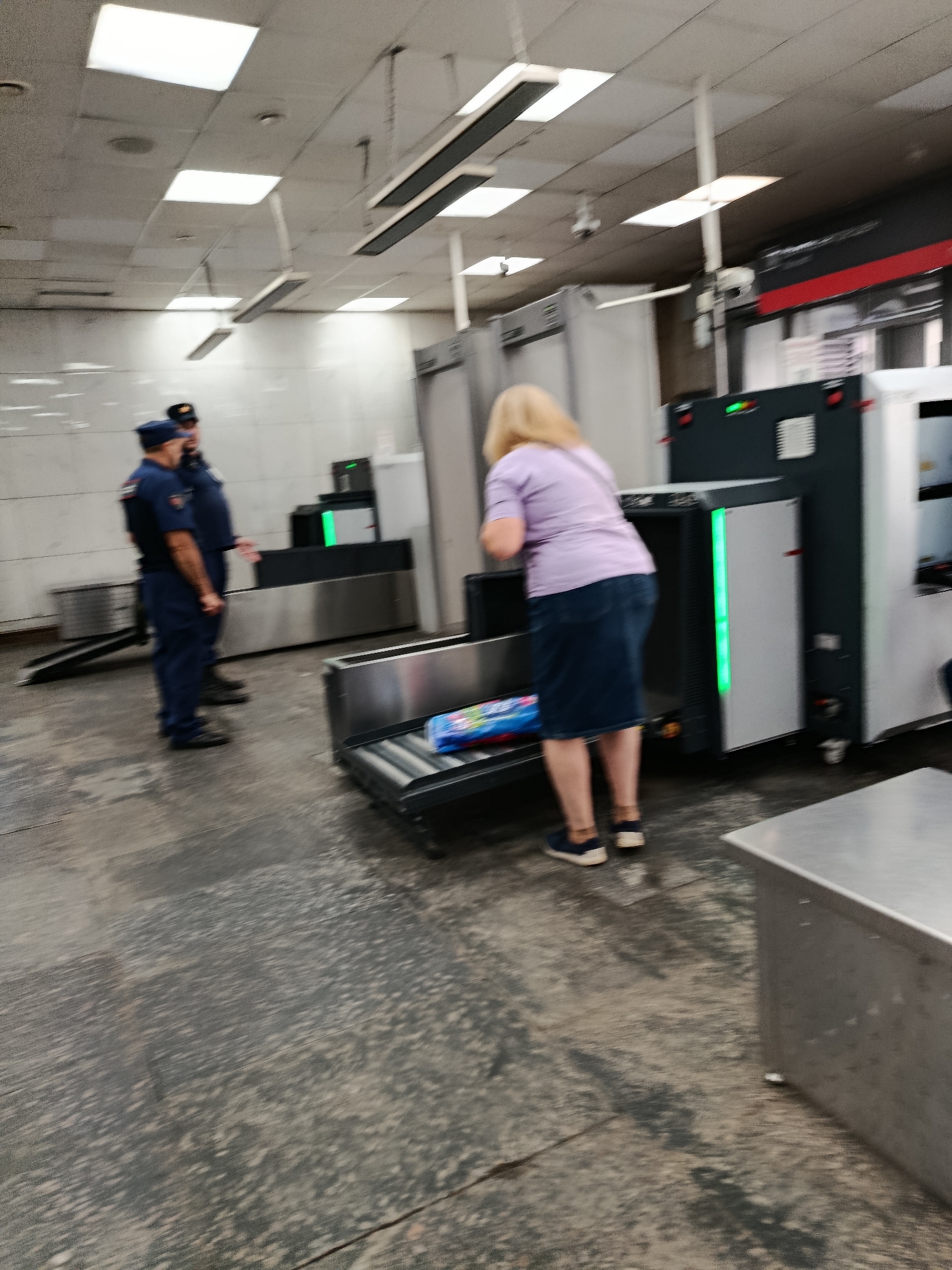 The image depicts a security checkpoint, likely at a Moscow Metro station, with two uniformed security guards and a woman placing an item into an X-ray scanner. The environment appears to be an indoor facility with fluorescent lighting, tiled walls, and a worn concrete floor. The security guards, dressed in dark blue uniforms with caps, stand near a metal detector archway, suggesting a controlled access point. The woman, wearing a purple shirt and a blue denim skirt, is the focus of the scanning area, interacting with the conveyor belt of the X-ray machine. The presence of security personnel and scanning equipment indicates a security screening process, common in public transportation hubs and important facilities within Moscow.