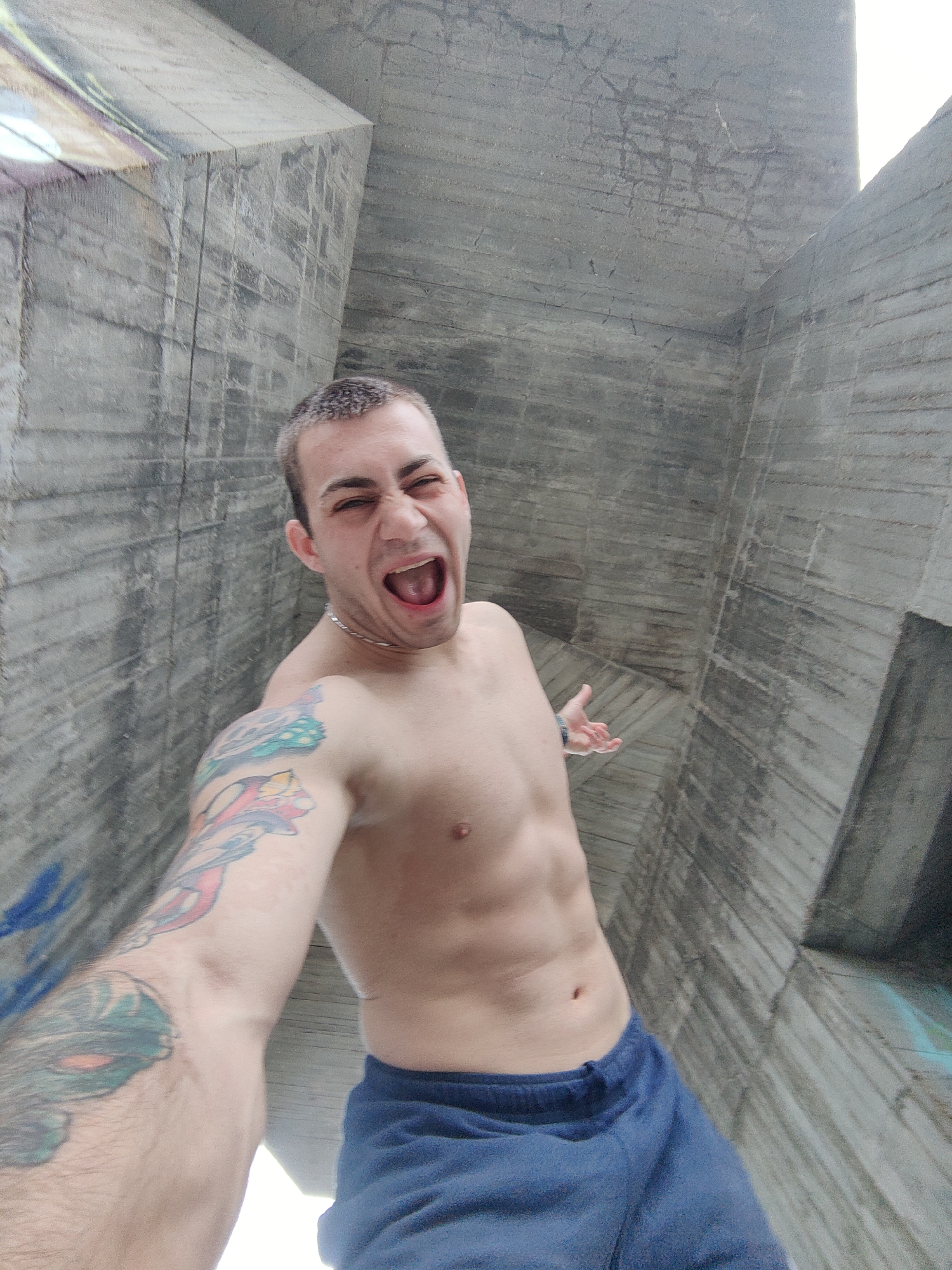 A shirtless man is taking a selfie in front of a concrete structure. He has a shaved head, a silver chain necklace, and tattoos on his left arm. He is wearing blue athletic shorts. His mouth is open and he is yelling. The concrete structure behind him is gray and appears to be part of a large building or architectural feature. There are traces of graffiti on the concrete. Based on the style of the concrete and graffiti, it is likely to be in Varna, Bulgaria.