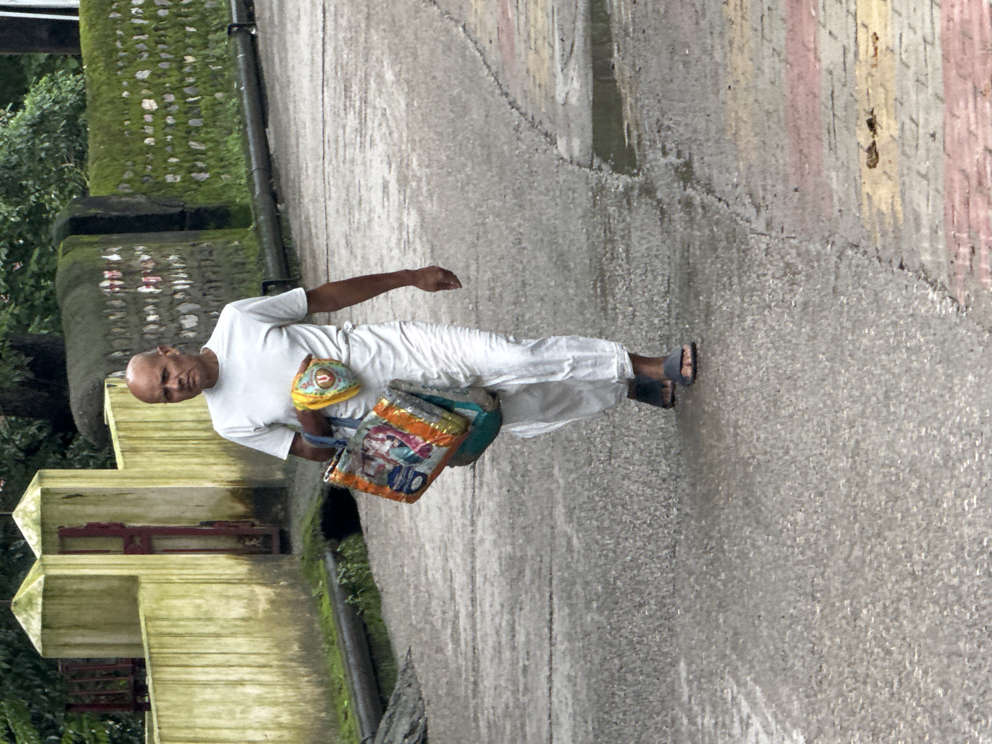 A bald, light-skinned man wearing a white t-shirt and white pants walks on a wet asphalt road in Rishikesh, India. He carries a colorful bag in his right arm. The road is bordered by a wall and some vegetation. The man appears to be walking towards the camera.