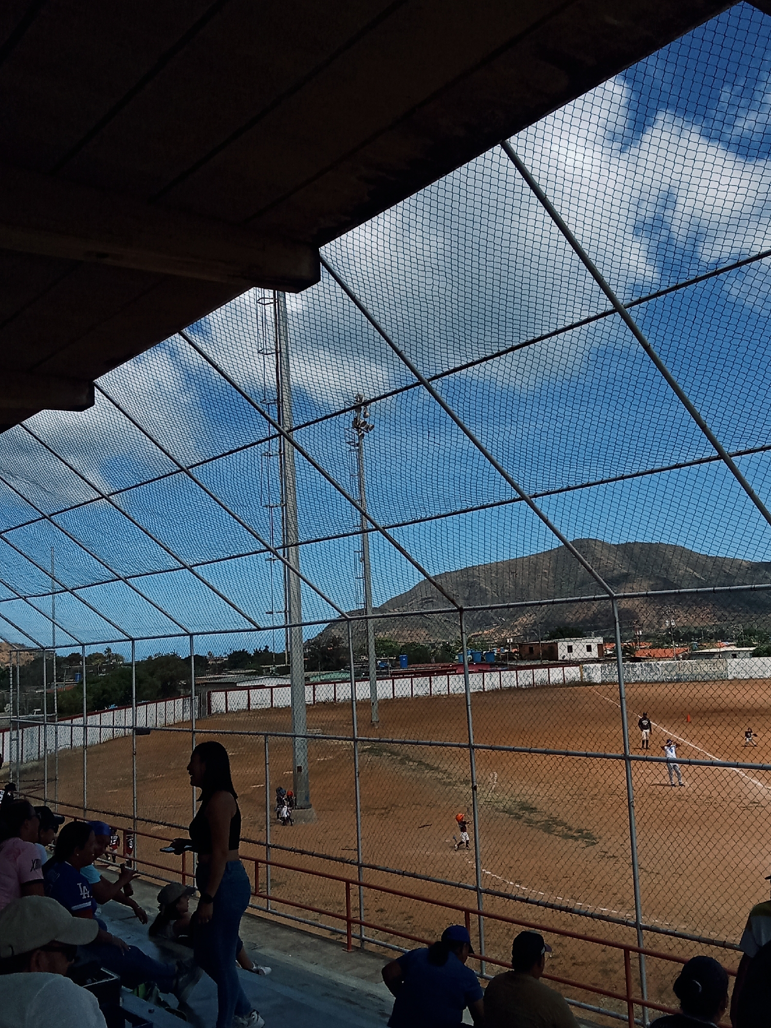 The image depicts a baseball game in progress, viewed from the stands of a stadium in Juan Griego, Venezuela. The field is a reddish-brown dirt, surrounded by a white wall and fence. A batter stands at home plate, a runner is on first base, and a fielder is positioned in the outfield. The stadium is enclosed by a high, wire mesh fence. The sky is blue with scattered clouds. The background shows a low mountain range and the buildings of Juan Griego. In the foreground, spectators are sitting and standing in the bleachers, including a woman in a black top and jeans standing in the aisle.