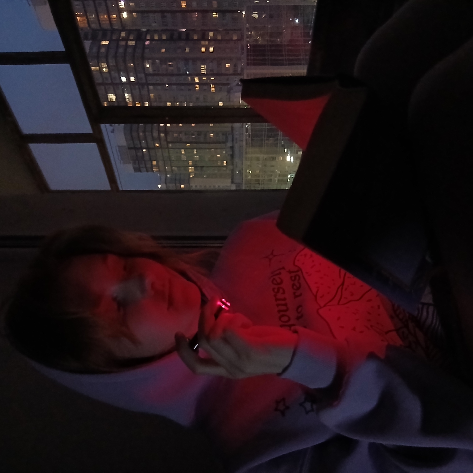 The image captures a person, likely female, indoors at night or late dusk in an urban setting, possibly Yekaterinburg, Russia, as suggested by the context. The individual is positioned beneath a large window, through which a cityscape with numerous illuminated high-rise buildings is visible. Reflections of these buildings are also present in the window panes.
The person is illuminated primarily by a strong red light emanating from an open book held close to them, casting a deep red glow on their face, hands, and a light-colored sweatshirt. Their face is partially obscured by shadows and the red light, but they appear to be looking down or away. In their hand, they hold a vaping device or electronic cigarette, which emits a small red light at its tip.
The person's sweatshirt features a graphic design with the partial text "yourself to rest" visible on the chest area, accompanied by star illustrations. The scene conveys a quiet and intimate atmosphere, likely an evening of reading and relaxation.