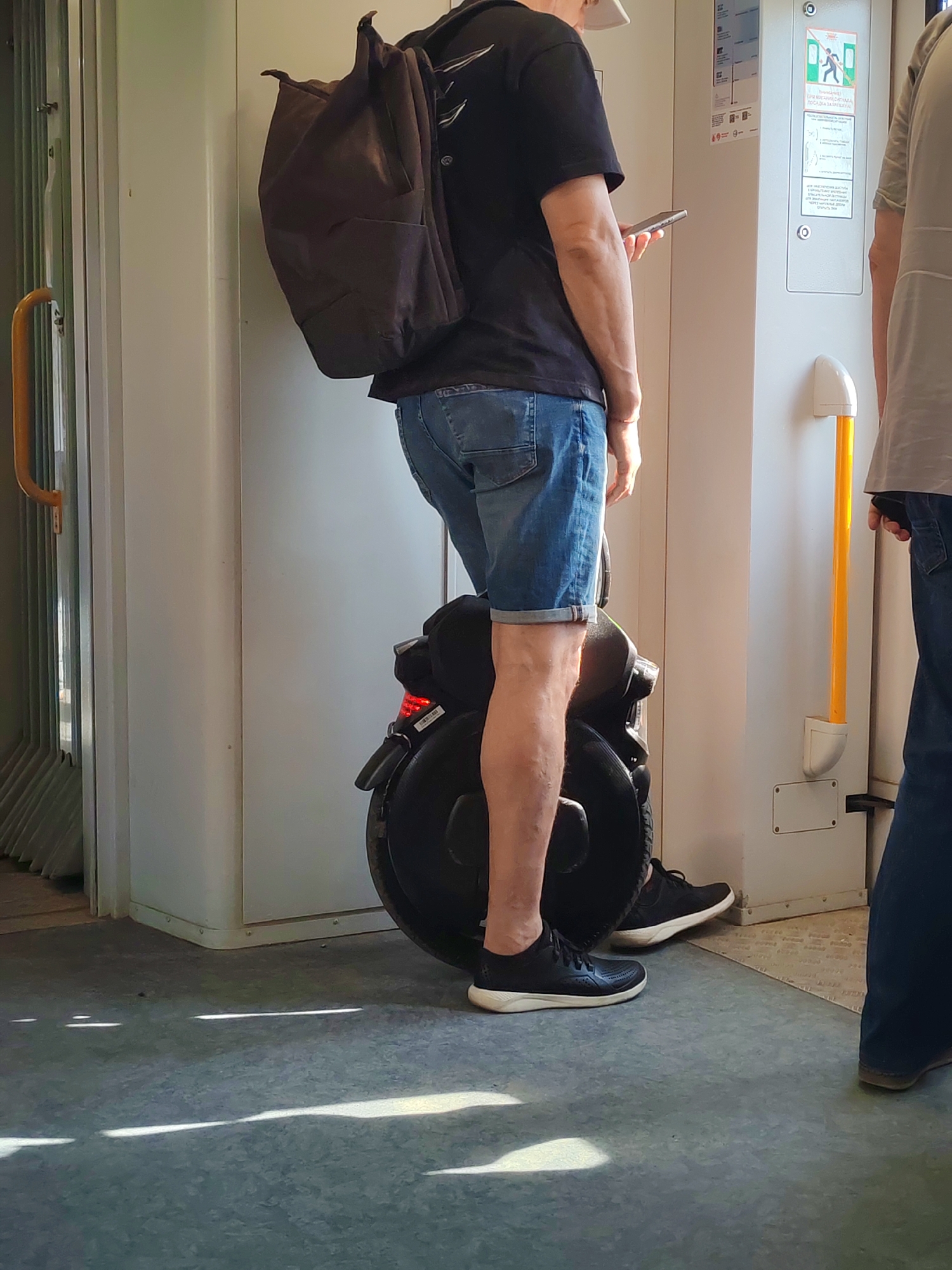 The image shows a man riding an electric unicycle inside a train car. He wears a black t-shirt, denim shorts, and dark sneakers. A brown backpack is slung over his shoulder. He holds a phone in his hand. To his right is another passenger in a grey shirt and jeans. The train car has white walls with informational signage and a yellow handrail. Light streaks across the grey floor. The location is Moscow, Russia.