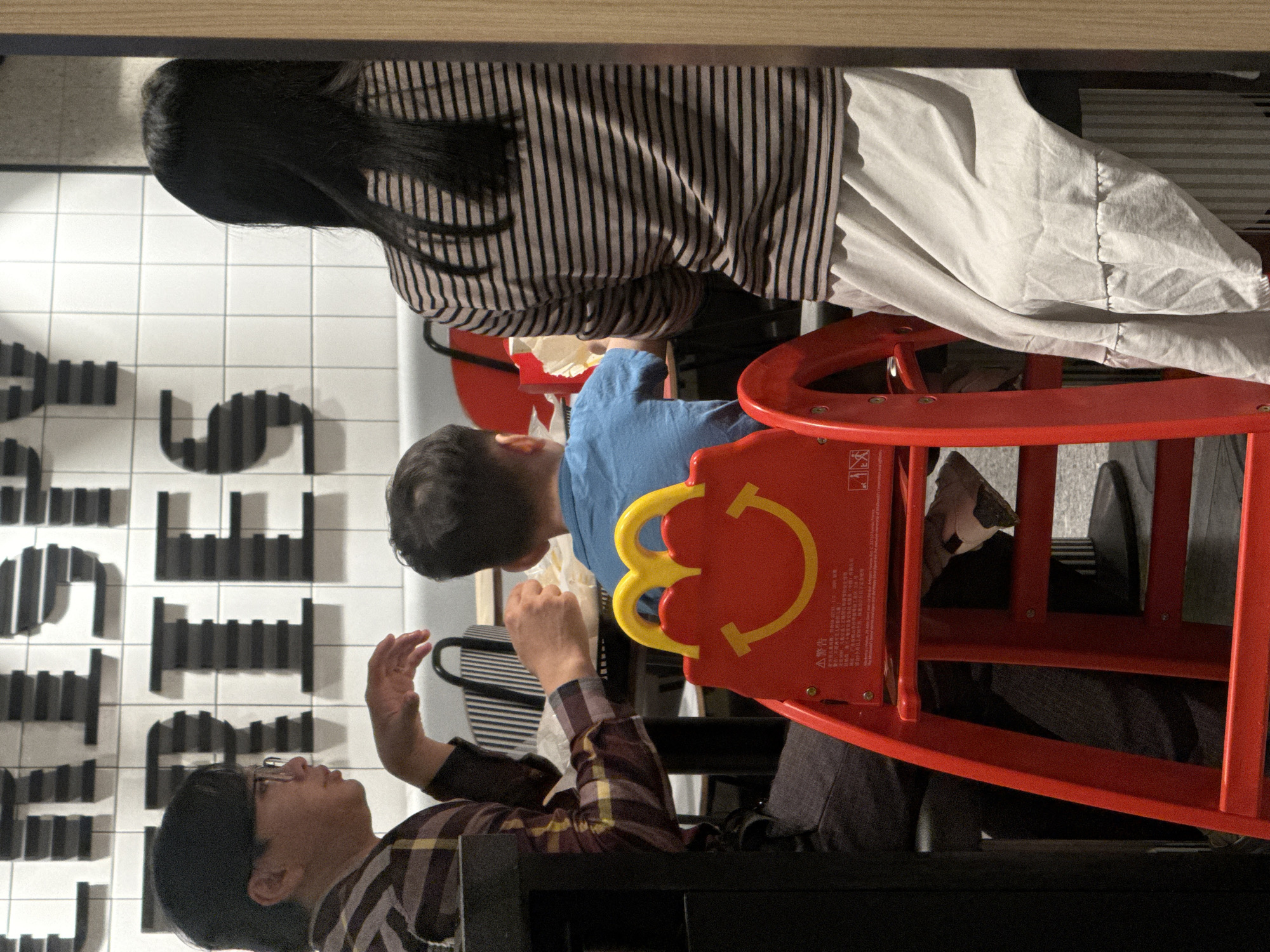In Da Lat, Vietnam, a family is seated at a table in a McDonald's restaurant. A man wearing glasses and a plaid shirt is on the left, talking with his hands. In the center, a young child in a blue shirt is facing away from the camera, playing with a red McDonald's Happy Meal toy slide. To the right, a woman with long dark hair, wearing a striped shirt and a light-colored skirt, is also seated at the table. The background features a tiled wall with large, dark lettering that spells out "CRISPY FRIES." The scene captures a casual family meal in a fast-food setting.