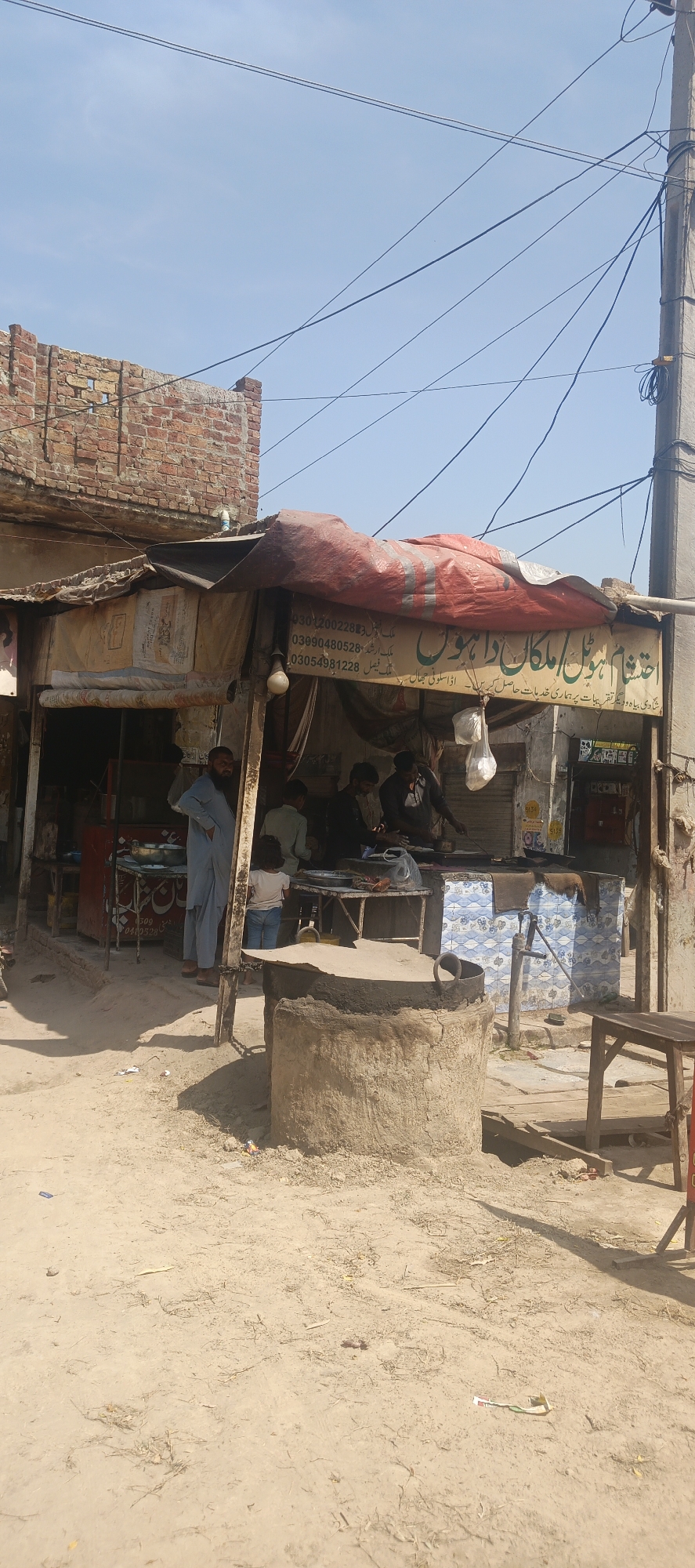 The image depicts an outdoor scene in Mandihar, Pakistan, likely a bustling street or market area during daytime under a clear sky. In the foreground, a large, round, mud-colored structure with a metal rim, possibly an outdoor cooking pit or a well, stands on unpaved ground. To its right, a simple wooden table is visible.
In the mid-ground, a makeshift food stall is set up, featuring a red tarp roof. Several men are present at the stall; one man in a light blue shalwar kameez stands prominently on the left, observing. Other men are actively engaged in preparing or serving food from various large pots and containers on tables within the stall. A small child is also seen near the stall. The ground in front of the stall is dirt, with some debris scattered.
Behind the stall, a weathered brick building with visible damage forms the backdrop. Overhead, numerous power lines crisscross the sky, attached to a prominent utility pole on the far right. A blue and white patterned tile structure, possibly a counter or a base for a water pump, is visible inside the stall area.
Visible text on the red tarp in Urdu includes contact numbers: "03012002288", "03090480528", "03054981228". Below the numbers, names like "ملک ارشد" (Malik Arshad) and "فیصل" (Faisal) are written. The main sign reads "اختشام ہوٹل امکان راہوں" (Ehtisham Hotel Imkan Rahoon), with a slogan below stating "شادی بیاہ و دیگر تقریبات پر ہماری خدمات حاصل کریں۔ اڈا سلونی جھال کو ملکی", which translates to "Our services are available for weddings, ceremonies, and other events. Adda Saloni Jhal Ko Malaki." The overall impression is that of a local eatery or catering service operating in a semi-urban or rural setting.