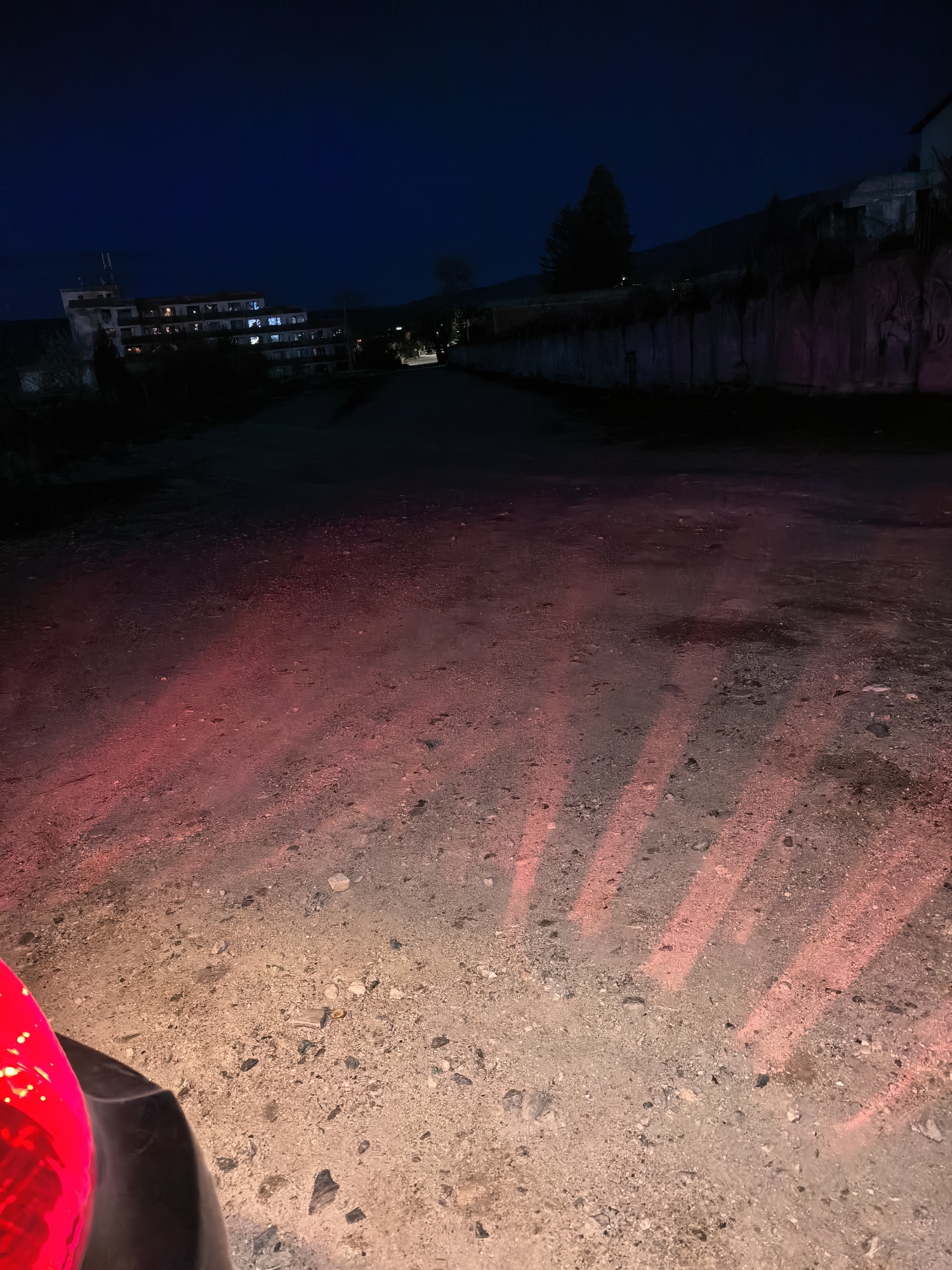 The image depicts a nighttime scene on a dirt road in an unknown city. The road leads into the distance, flanked on one side by a wall and on the other by buildings with illuminated windows. The foreground of the road is illuminated by a red light source from an unseen object. The sky is dark. The road appears unpaved and gravelly. There are no people visible.