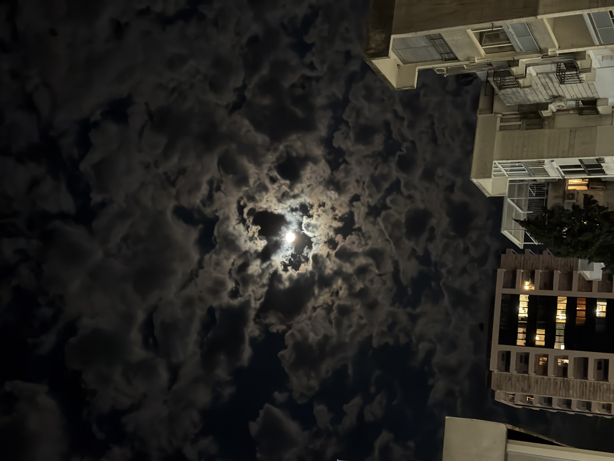 The image captures a dramatic night sky with a prominent moon, viewed from an upward angle, framed by urban architecture. The scene is dominated by a dark blue-black sky filled with numerous, textured clouds. The moon, bright and nearly full, is centrally located and partially obscured by these clouds, creating a luminous glow that highlights their contours and gives them a dynamic, almost swirling appearance.
On the right side of the frame, parts of several multi-story buildings are visible, suggesting an urban or suburban setting. These structures are light-colored, possibly concrete or stucco, featuring multiple windows, some of which are illuminated from within, indicating human presence. Balconies with railings and what appear to be air conditioning units are also discernible on the building facades. A small patch of dark green foliage, likely a tree, is visible near the bottom right. No people or specific activities are depicted, and no visible text is present. The time of day is clearly night, with the moon being the primary light source illuminating the clouds and casting faint light on the upper parts of the buildings. The perspective is from below, looking up towards the sky and the surrounding structures.