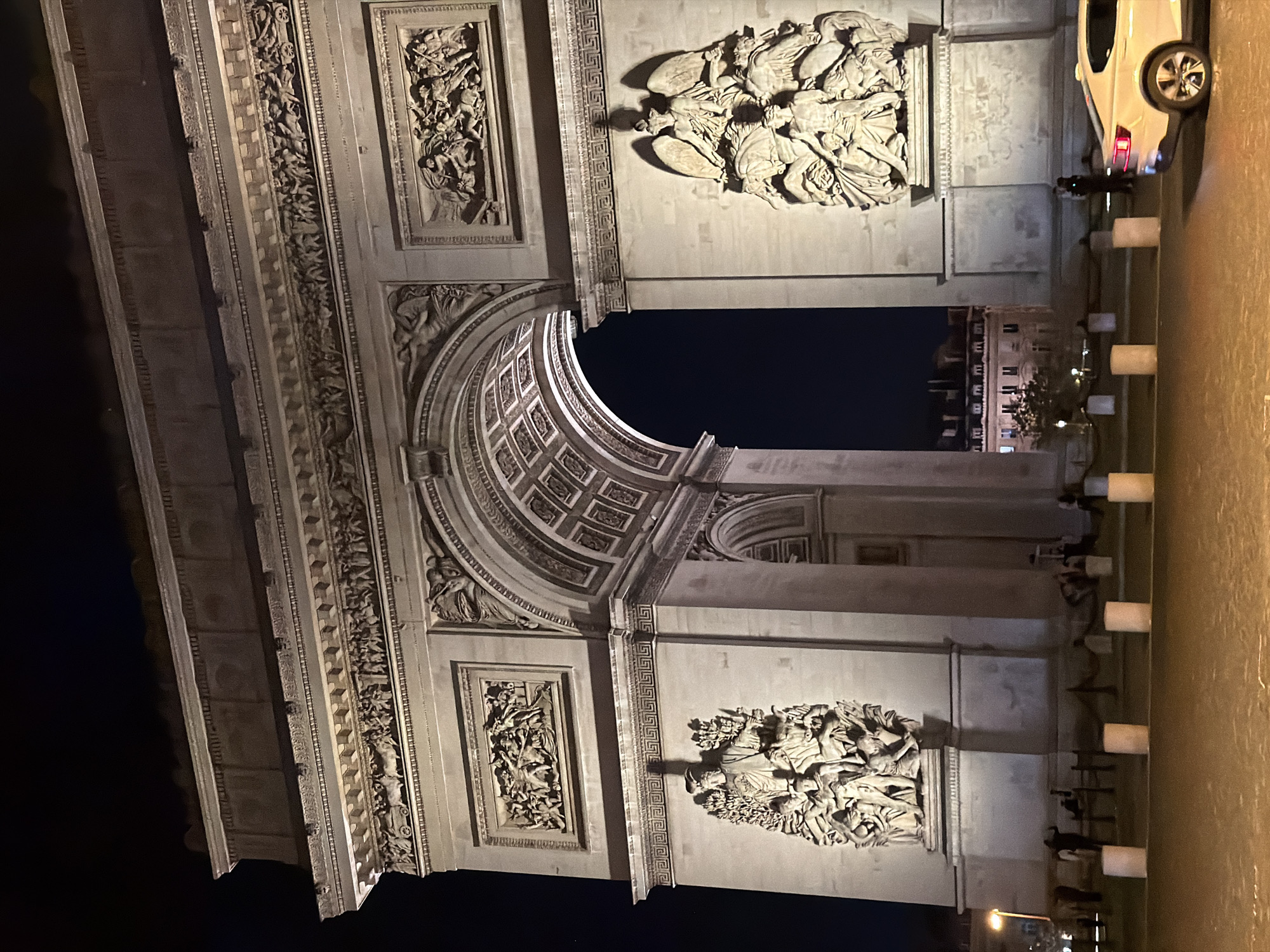 The image captures the Arc de Triomphe at night in Paris, France. The architectural details, including sculptures and moldings, are illuminated. People are visible near the base of the monument along with bollards and parked car on the right. Another building can be seen through the archway. The sky is dark.