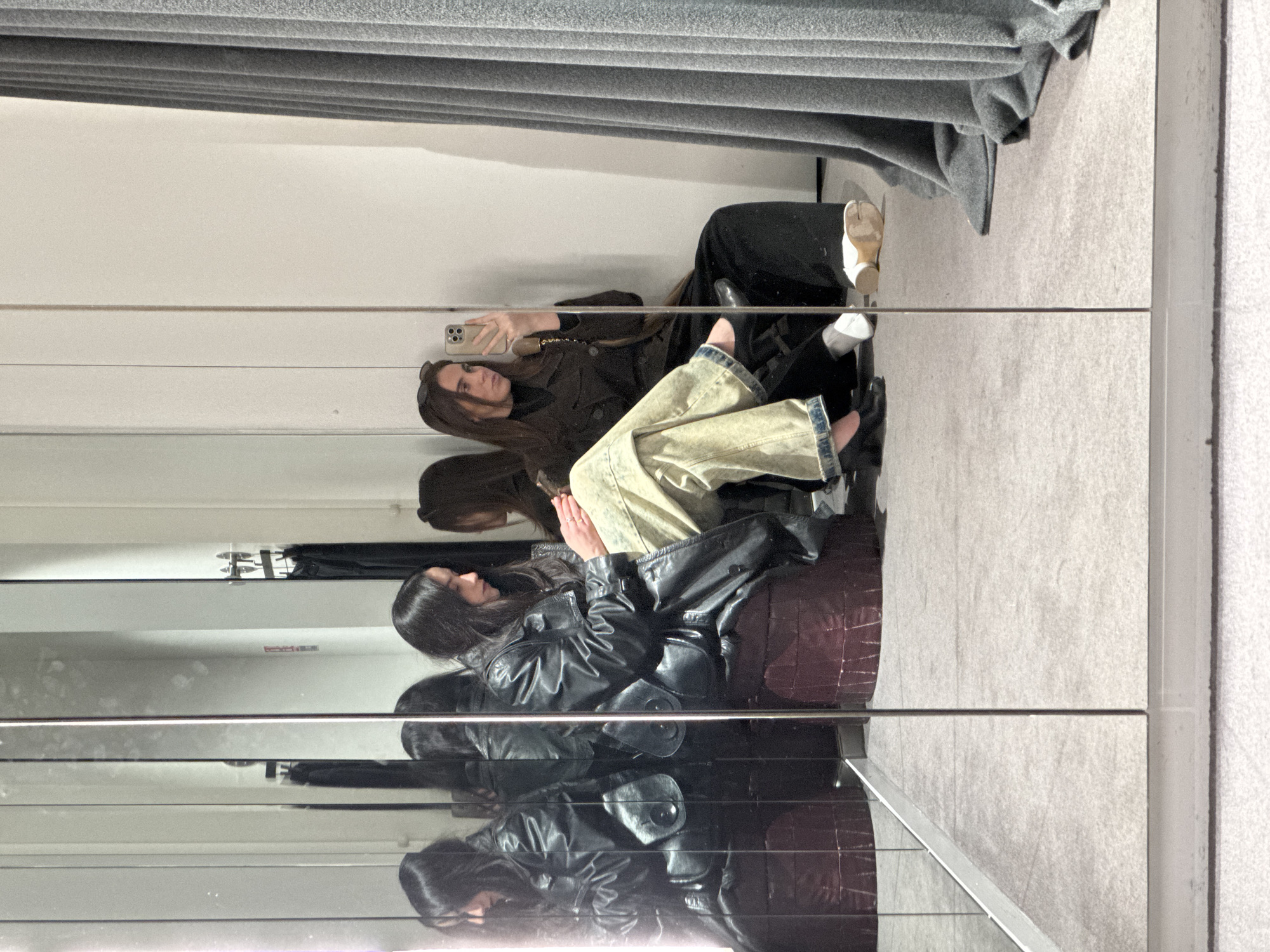 The image captures a scene reflected in a mirror, likely in a retail dressing room. Two women are present. One woman is seated, wearing a dark jacket, light-colored jeans, and dark shoes. She is holding a mobile phone and appearing to take a picture or selfie. The second woman is seated next to her, wearing a dark leather jacket and dark trousers. Clothing racks with grey curtains can be seen at the top of the image. The location may be in Paris, France, but this is not evident from the image itself.