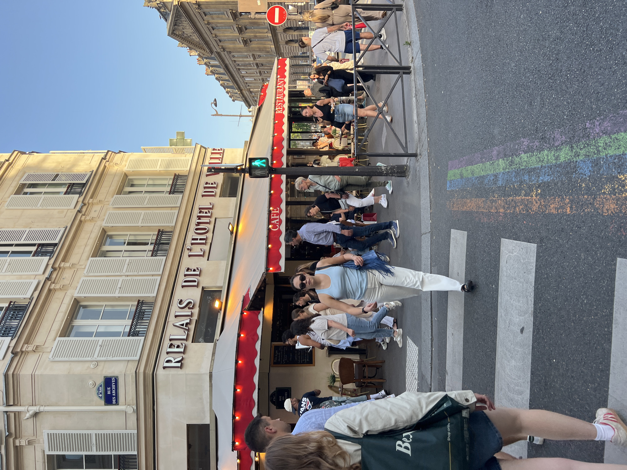 The image captures a street scene in Paris, France, specifically at the "Relais de l'Hotel de Ville" cafe and restaurant located on Rue des Archives in the 4th arrondissement. A group of pedestrians is crossing the street at a zebra crossing, with a woman in a light blue top and white pants prominently in the foreground. The cafe's red and white awning extends over an outdoor seating area, where people are sitting. A traffic light shows a green "walk" signal. A no-entry road sign is visible near a metal guardrail. Rainbow-colored stripes are painted on the road surface next to the zebra crossing. The building adjacent to the restaurant has light-colored stone and white shutters on the windows.