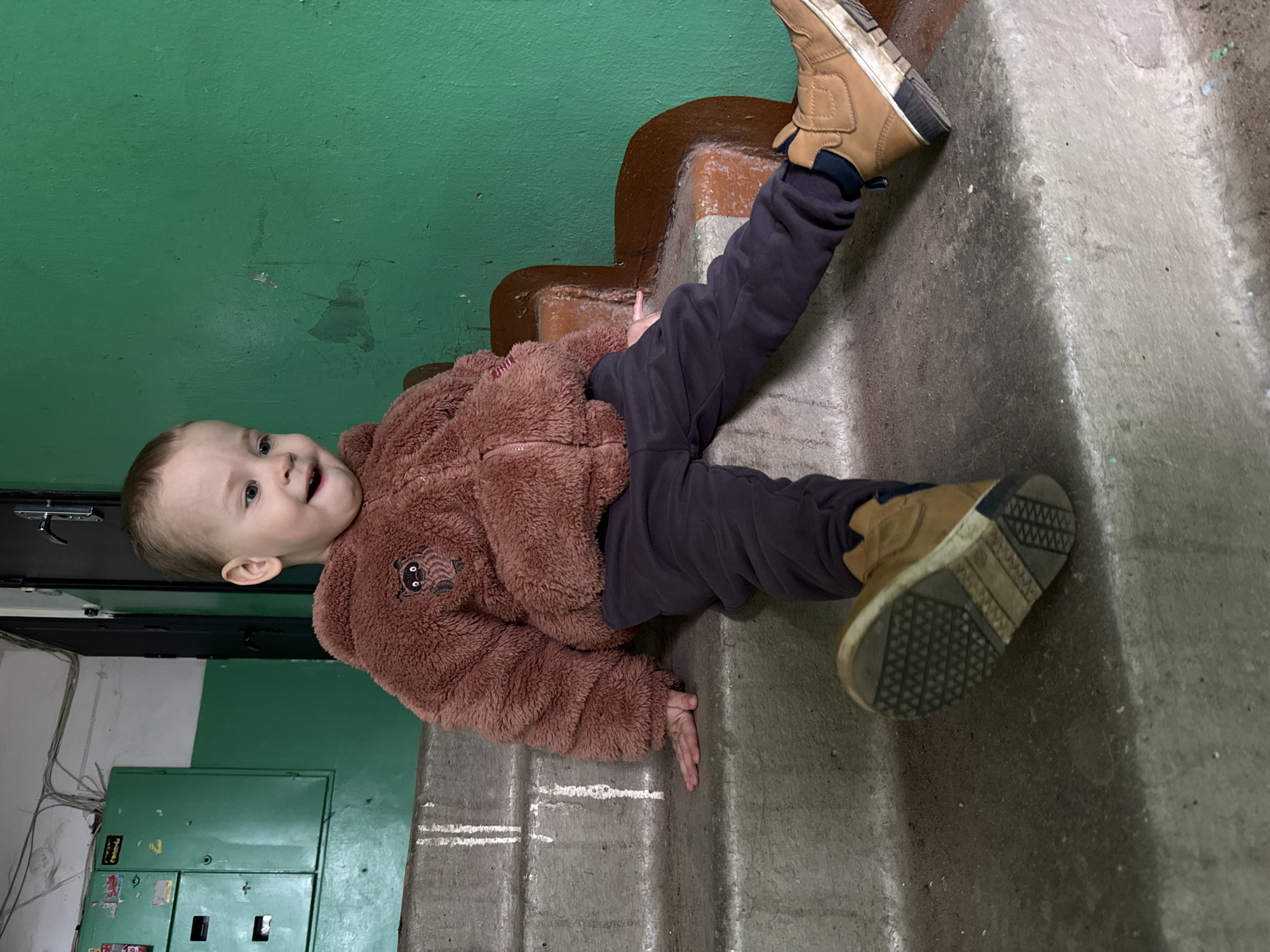 A young child wearing a brown, furry jacket and dark-colored pants is sitting on concrete stairs. The child is also wearing brown boots. The stairs are inside, near a green wall, possibly in an apartment building. An electrical panel is visible on the wall. The location appears to be an urban area within a city, but the specific city and state are unknown.