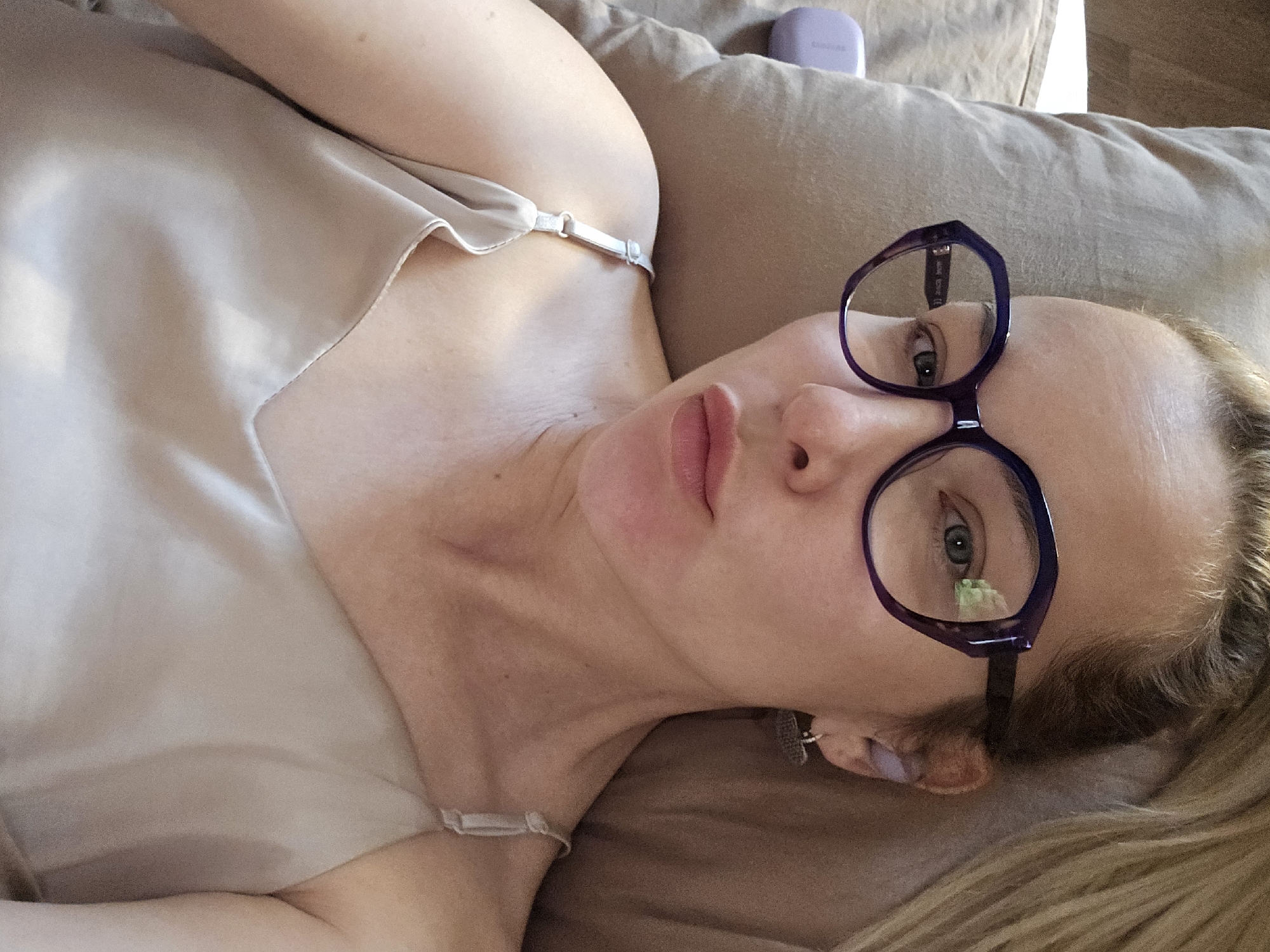 A woman is lying on a beige surface, possibly a sofa or bed. She is wearing a light-colored silk or satin top with thin straps and large, purple-framed glasses. The woman has fair skin and light-colored hair. A small, light purple electronic device is visible in the upper right corner of the image. The scene appears to be indoors. There is no indication that the image was taken in Sochi, Russia.