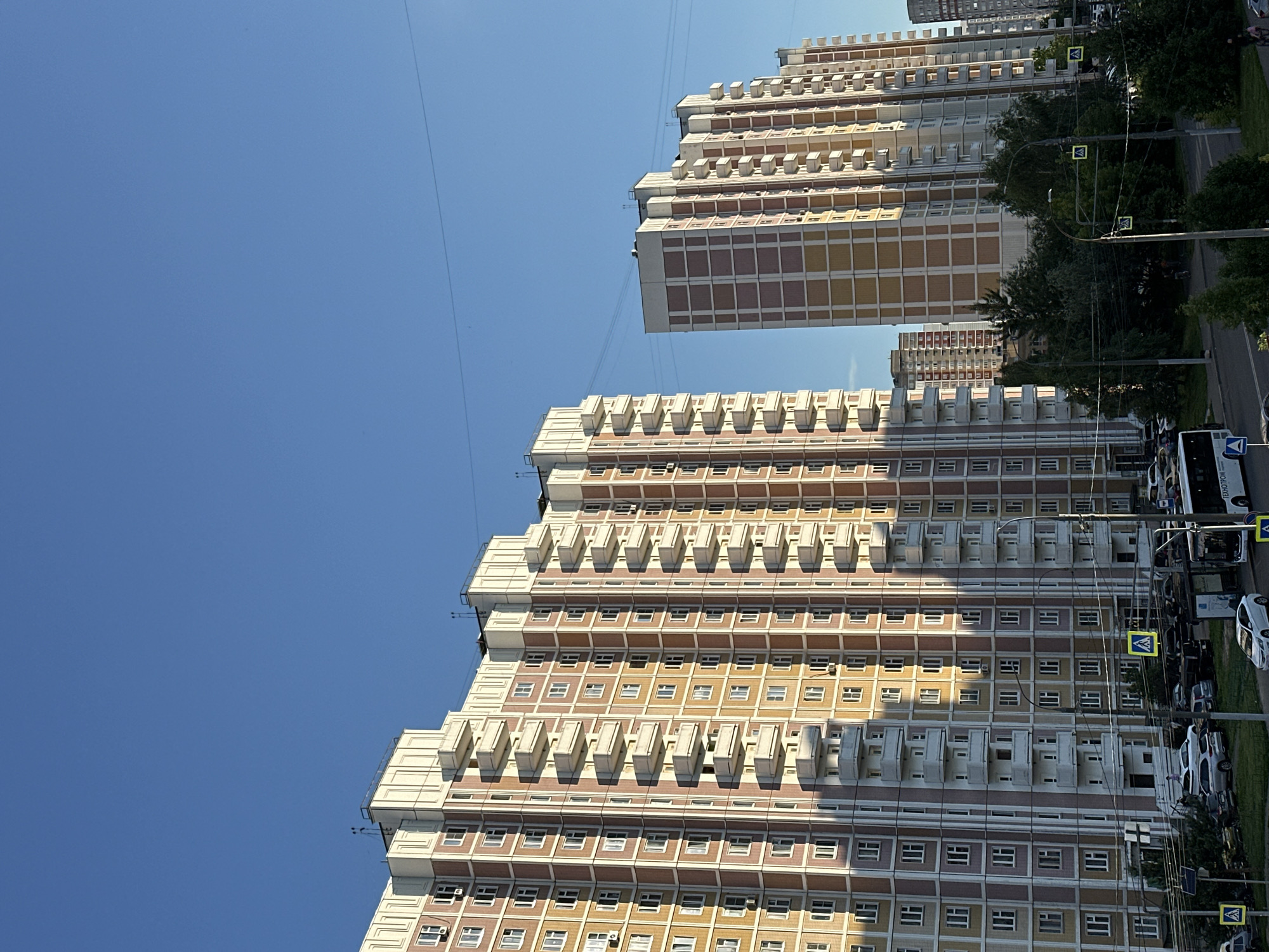 This media file captures a clear, sunny day in Podolsk, Russia, with tall, residential buildings dominating the scene. The sky is a vibrant, cloudless blue. The buildings are multi-story apartment complexes, characterized by repetitive modular balconies and numerous windows. The facade of the buildings features a color palette of beige, yellow, and reddish-brown square tiles, creating a visually distinct pattern.
In the foreground, the lower portion of a building shows more detailed architectural elements, including visible air conditioning units on some balconies and a row of windows with white frames. In the background, another similar building stands to the left, also showcasing its repeating balcony and window design. The buildings are juxtaposed against the bright sky, with power lines subtly stretching across the frame.
To the right of the buildings, a street is visible with parked cars and a white bus. Several yellow pedestrian crossing signs are mounted on poles along the street, indicating a residential area. Lush green trees and grass line the street, adding a touch of nature to the urban landscape. The lighting suggests it is daytime, likely late morning or early afternoon, given the strong sunlight casting shadows from the balconies. There is no discernible activity of people in the frame, but the presence of vehicles and pedestrian crossings implies a populated, functional neighborhood. One visible text element on the bus reads "ПОДОЛЬСК" (Podolsk), confirming the location.