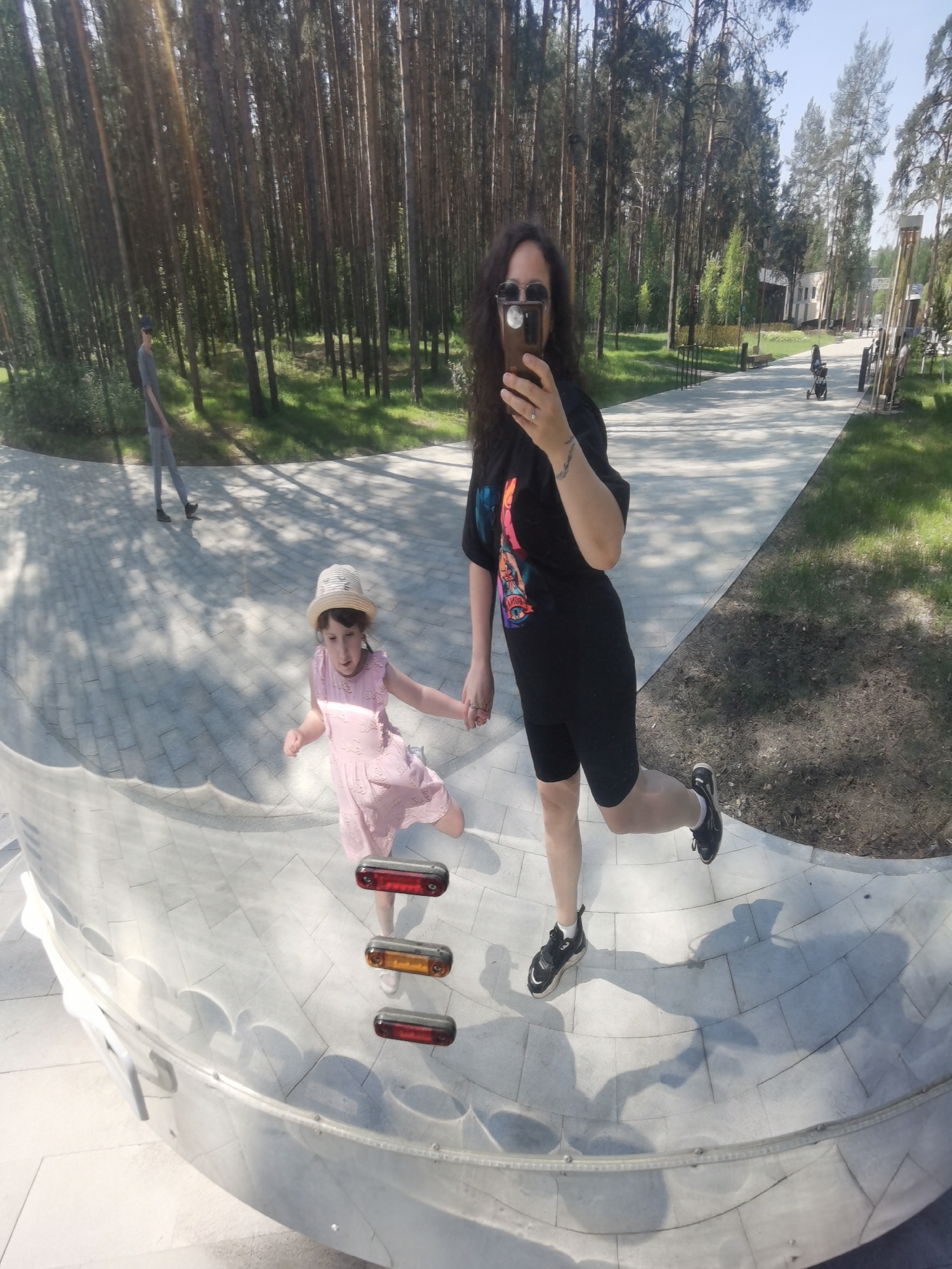 The image depicts a woman holding a cell phone, likely taking a photo, standing in a park in Perm, Russia. She is holding the hand of a young girl in a light pink dress and a hat. They are standing next to a curved, mirrored structure which distorts the reflection of the surroundings. The background shows a paved walkway, grass, and tall trees. In the distance, there are other people and buildings.