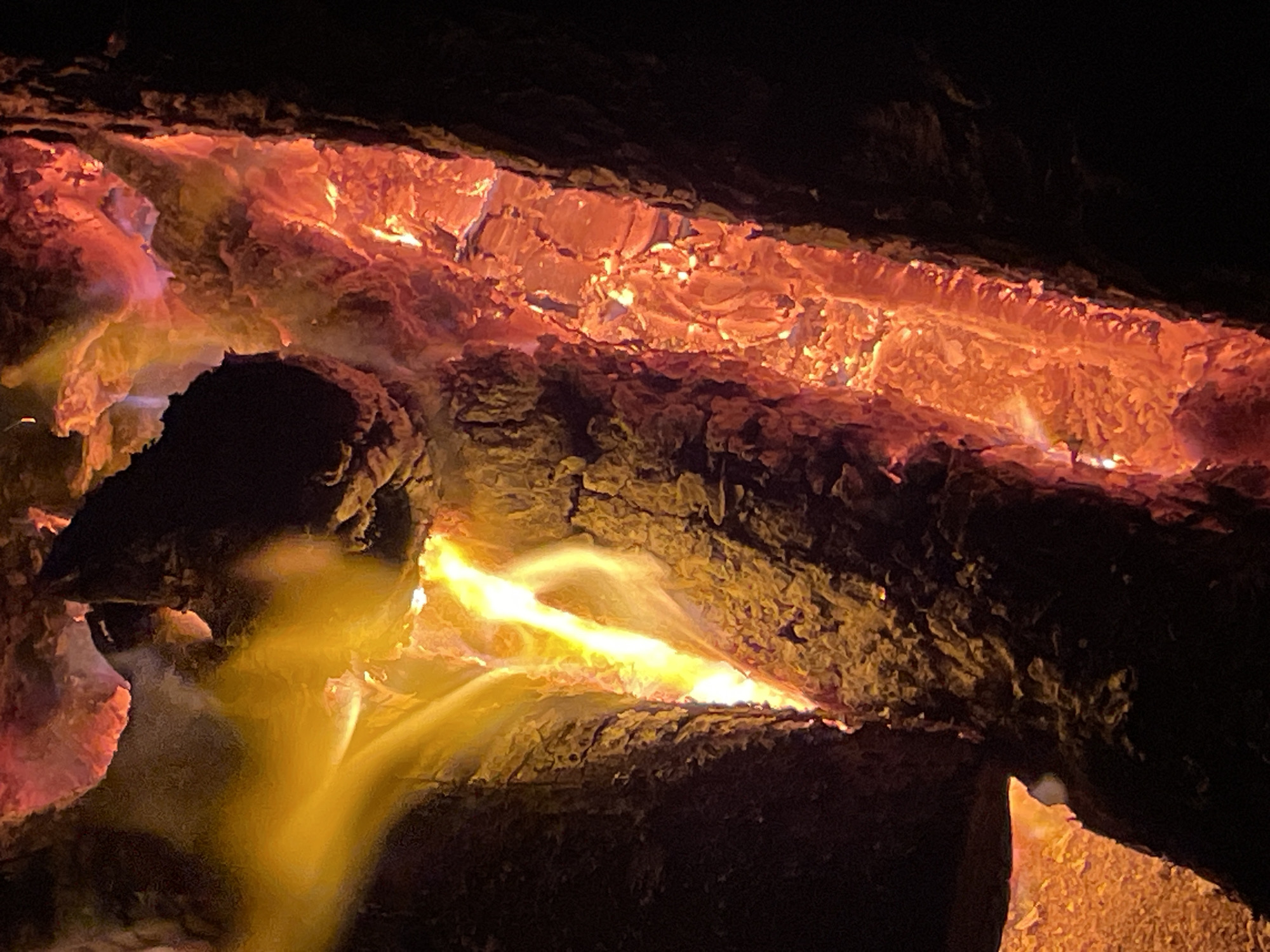 This image presents a close-up, vertical view of burning wood, likely within a campfire or fireplace, located in the city of Zelenaya Roshcha, Russia. The scene is illuminated by the intense heat and light generated by the combustion.
The primary subjects are several pieces of wood in various stages of burning. On the left, a large, dark, irregularly shaped piece of wood displays bright yellow and white flames ascending in a prominent, luminous streak towards the upper-left quadrant. The wood surrounding these flames is glowing with deep orange and red embers, revealing a rough, textured surface.
Towards the center and extending to the right, another substantial piece of wood is intensely incandescent, glowing bright red and orange. This section exhibits a highly irregular, textured surface with visible cracks and crevices filled with glowing material, suggesting active charring and ember formation.
A distinct, vertical band of intensely glowing, fragmented material, consistent with burning wood and embers, runs along the right side of the image. This band is predominantly red and orange, with numerous brighter hot spots, and contrasts sharply with the unlit, dark background on the extreme right. No human figures or discernible objects other than the burning wood are present in the frame.