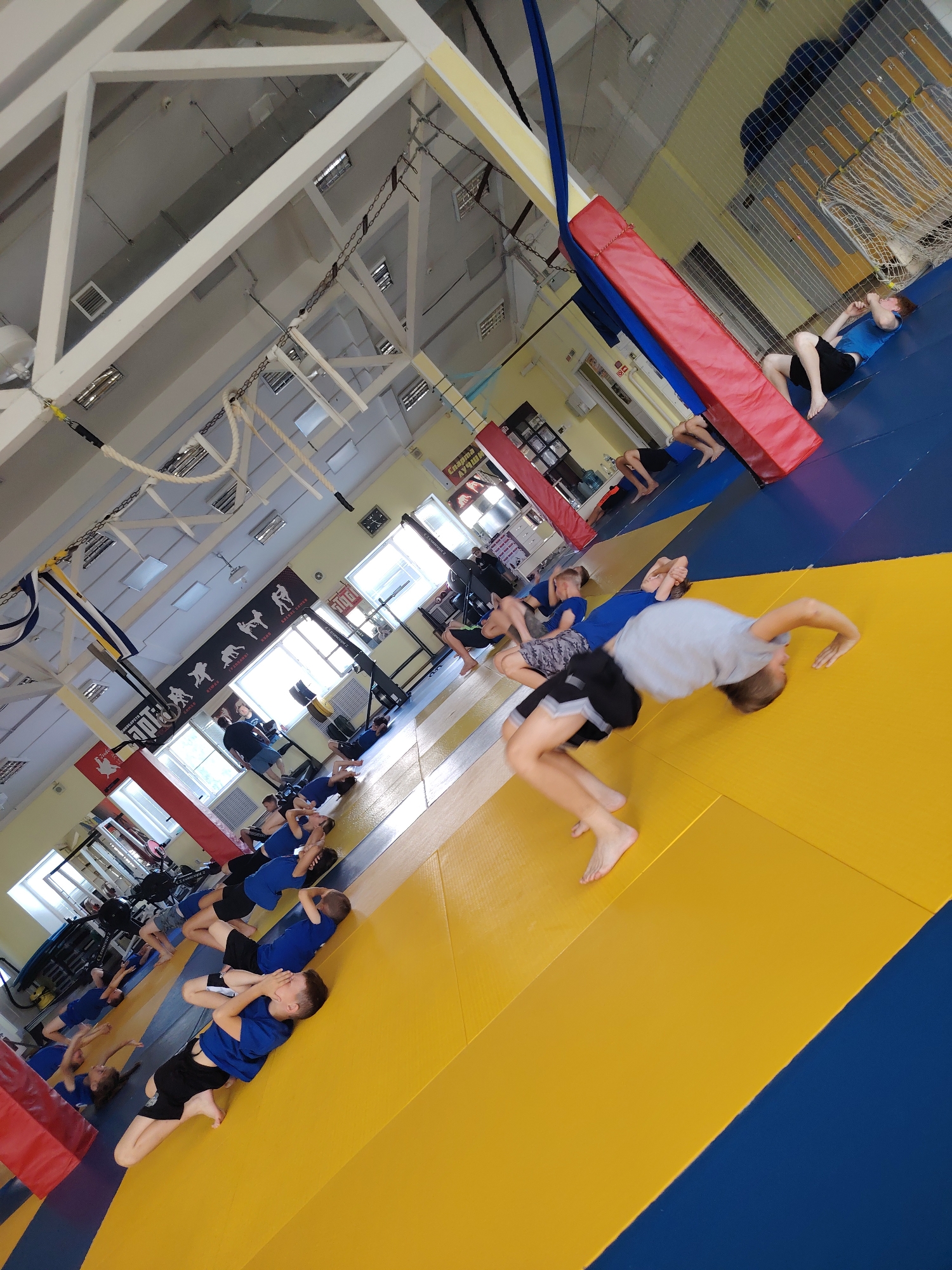 The image depicts an indoor gymnasium in Chaikovsky, Russia, likely a wrestling or martial arts facility, with blue and yellow mats covering the floor. Several individuals, predominantly young boys and a few young girls, are engaged in physical exercises. Some are lying on their backs with hands behind their heads, others are in a seated position, and one boy in the foreground is captured mid-somersault. In the background, a person is seen lying down near a red padded obstacle. The gym appears to have a high ceiling with exposed beams and industrial lighting. There are also various pieces of gym equipment visible, including what appears to be rowing machines and weight racks. Banners with silhouettes of athletes in action are displayed on the walls. The overall scene suggests a training session or practice.