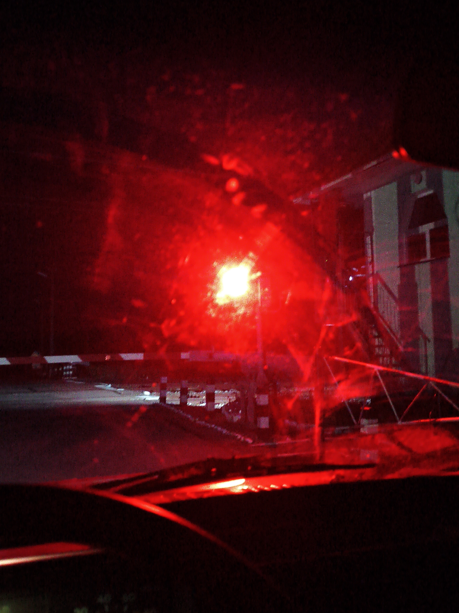 The image depicts a nighttime scene with prominent red lighting, suggestive of a hazard or warning signal. In the foreground, a dark car interior is visible, with a portion of the windshield and dashboard in shadow. The windshield appears to have some water droplets or smudges.
Beyond the car, a road or pathway is illuminated by the intense red light, which also reflects off a surface in front of the vehicle. A barrier, possibly a railway crossing gate, with alternating white and red stripes, is visible across the road. Behind the barrier, a small building or structure with a striped facade and a window is partially visible on the right side of the frame. The overall atmosphere is dark and somewhat distorted by the bright red light and the condition of the windshield. The location is identified as Hoholeve, Ukraine.