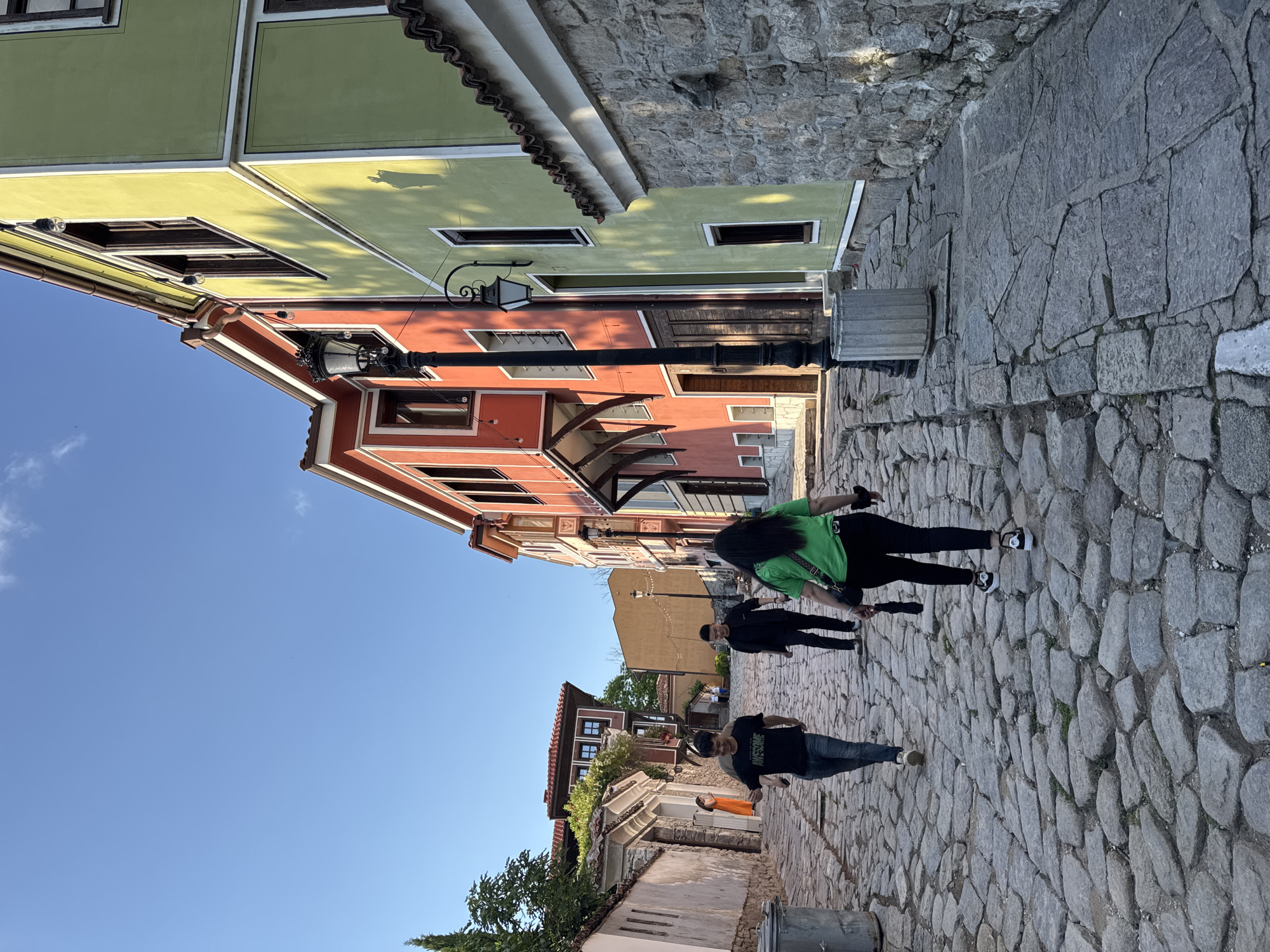 The image captures a street scene in Plovdiv, Bulgaria, characterized by cobblestone paving and colorful, traditional buildings lining the street. Three people are walking along the street. The person closest to the camera wears a black t-shirt and jeans. Another person in black clothing walks in the middle. The person furthest from the camera wears a bright green t-shirt and black pants. The buildings are painted in various colors, including yellow, orange, and red. A light fixture is seen extending from a red building. The sky is clear and blue. A stone wall borders one side of the street.