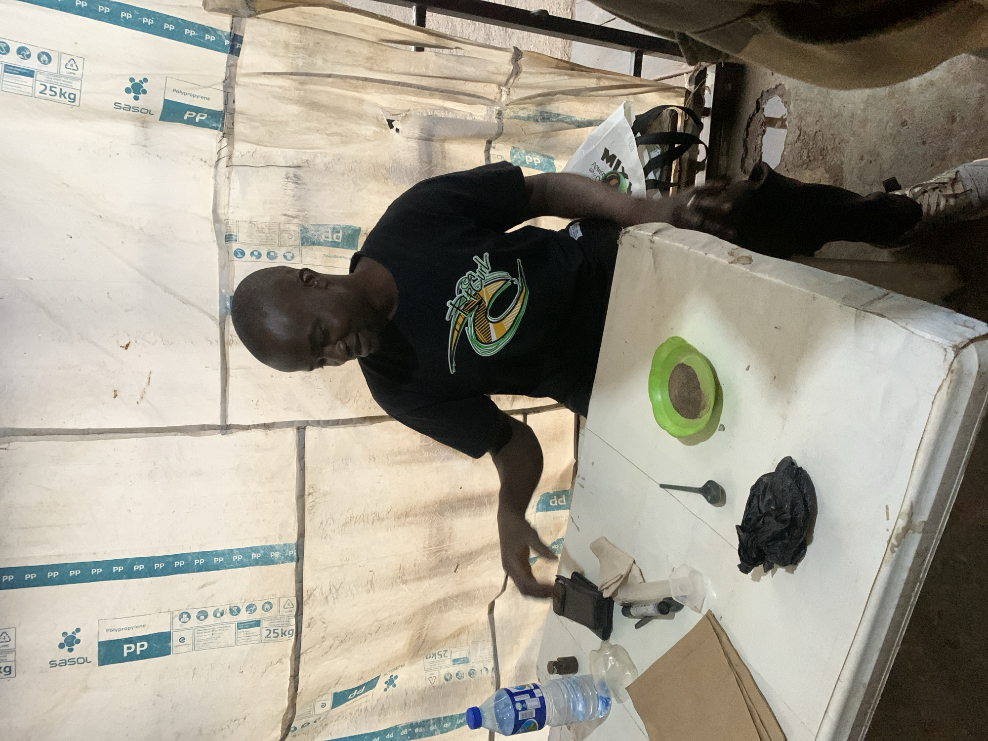 A black man wearing a black t-shirt with a circular green and yellow logo is seated at a white table in what appears to be an outdoor or semi-enclosed setting. The background consists of a taut, white, tarp-like material with prominent blue markings that read "SASOL PP 25KG". The man is looking down and to his right, with his left hand resting on the table near a black object. On the table are several items, including a plastic water bottle, a small dark vial, a syringe, a crumpled black plastic bag, a small black spoon, and a green plastic bowl containing a brown powder. A brown paper bag is also on the table to the left. The overall environment suggests a makeshift workspace or a temporary setup, potentially in an urban or semi-urban area of Jos, Nigeria. The ground beneath the table is rough concrete.