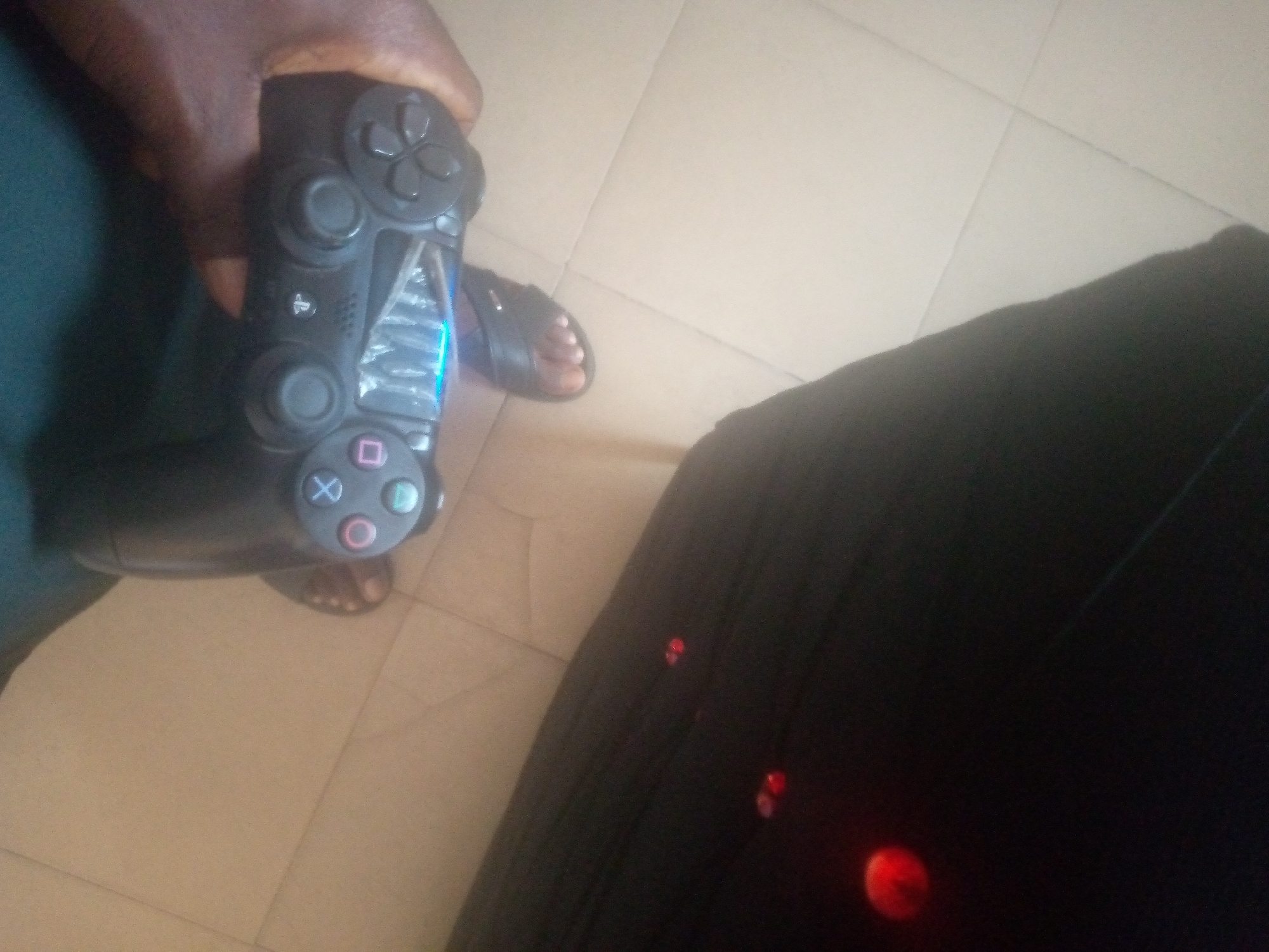 This image, described as being taken in Jalingo, Nigeria, captures an overhead view of a person's hand holding a black PlayStation DualShock 4 controller. The controller is actively emitting a bright blue light from a section near its center, which appears to be cracked or damaged. Below the controller, a dark-skinned foot, clad in a black slip-on sandal, rests on a light-colored tiled floor with discernible grout lines. To the right, a significant portion of a dark, possibly black, fabric or surface is visible, adorned with several scattered red, circular reflections or light sources of varying sizes and intensities. A dark green or teal garment is partially visible under the arm holding the controller.