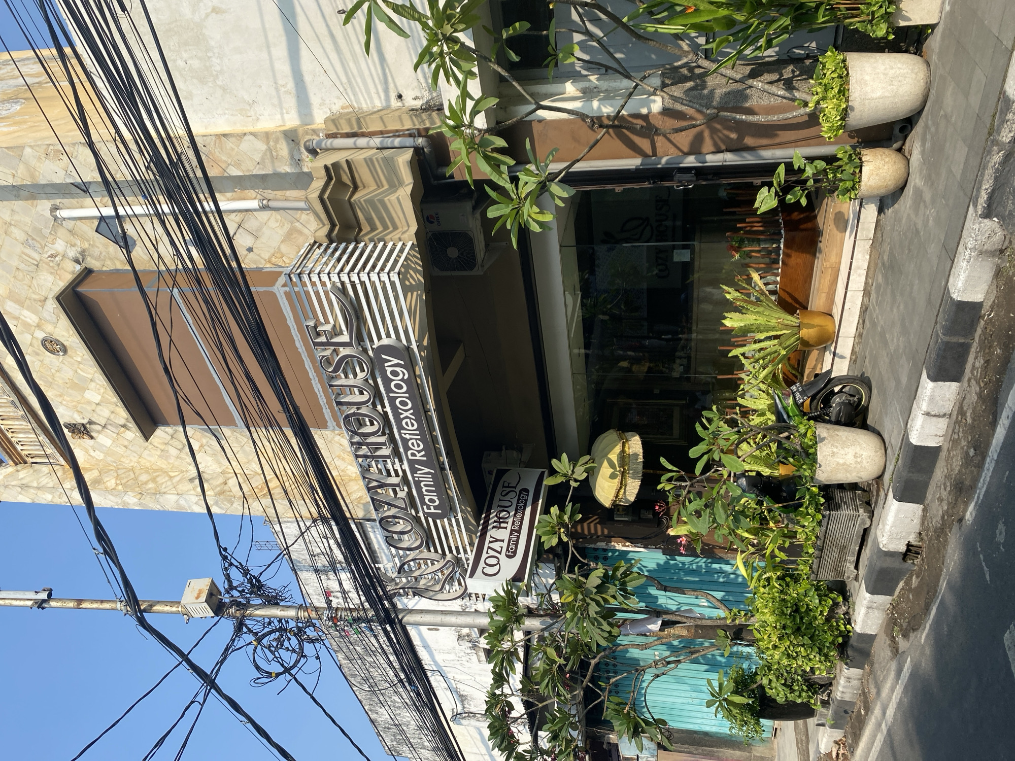 The image depicts the exterior of a business called "Cozy House Family Reflexology" located on a street in Kediri City, Indonesia. The building features a stone facade and a prominent sign with the business name. A variety of potted plants are arranged along the sidewalk in front of the establishment, contributing to a green and somewhat tropical aesthetic. Overhead, a dense network of electrical wires and a utility pole are visible against a clear blue sky, suggesting an urban setting with extensive utility infrastructure. A motorcycle is parked near the entrance, and a blue metal door is visible to the left of the building. The scene is captured during daylight hours, with shadows indicating the sun's position.