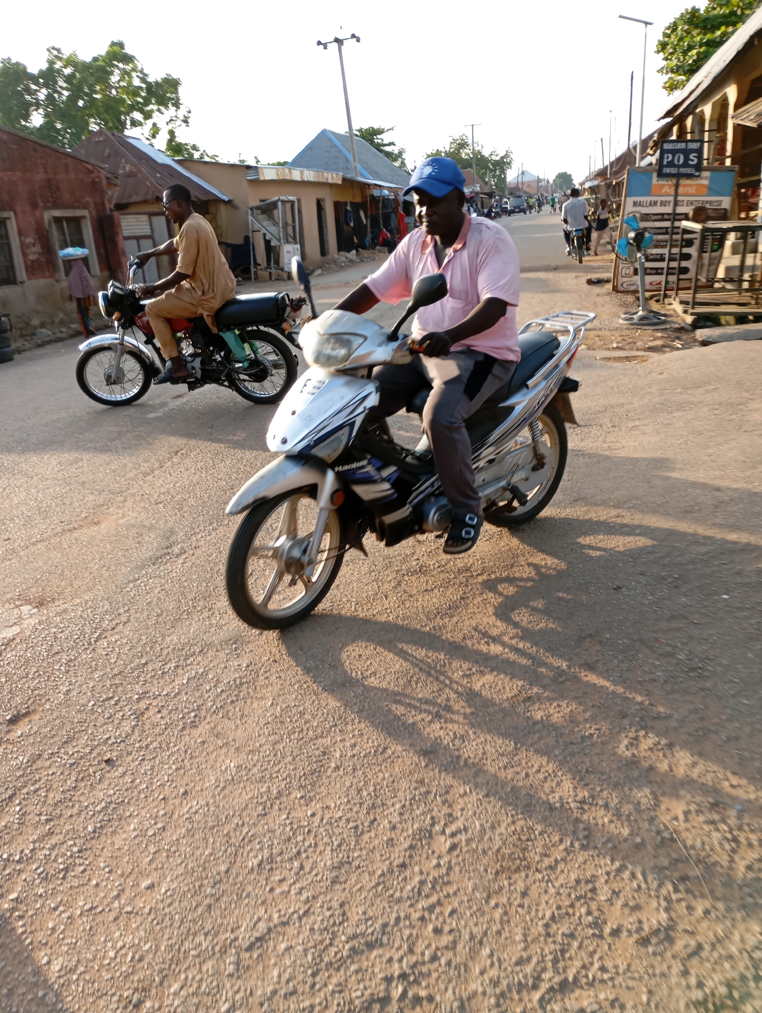 Two men ride motorcycles on a dusty, unpaved road in Shendam, Nigeria. The man in the foreground, wearing a pink shirt, blue cap, and gray pants, is on a silver motorcycle and is facing slightly to his left. Behind him, another man in a tan outfit rides a red and black motorcycle, also facing slightly to his left. The background shows a street lined with low-rise buildings, some appearing to be shops, with signs indicating services such as "POS" and "ENTERPRISE". The lighting suggests it is late afternoon, with long shadows cast across the road. The environment is a typical street scene in a Nigerian town.