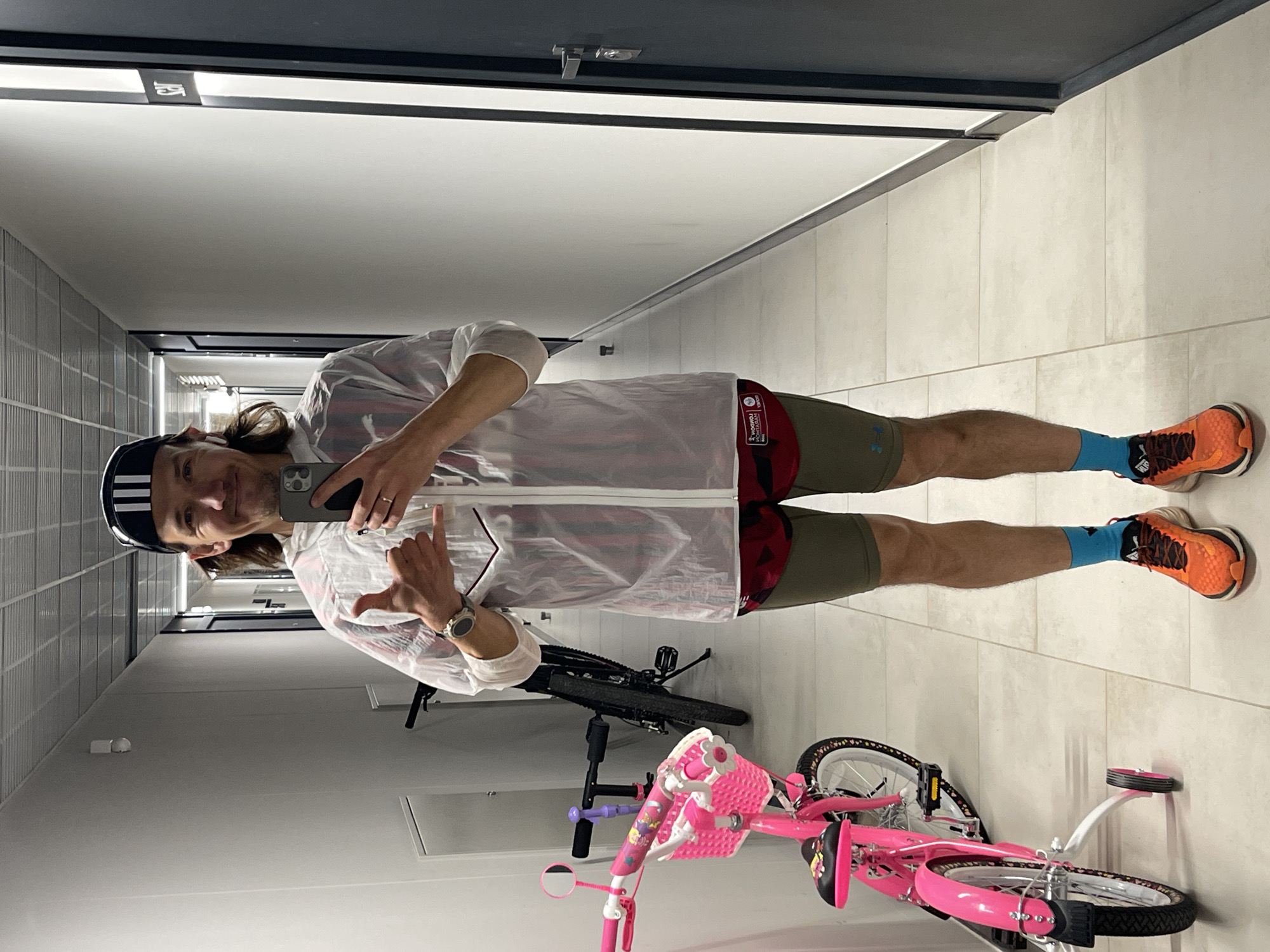 A man stands in a hallway, taking a selfie. He wears a black and white striped cap, a transparent white jacket, green shorts, and bright orange running shoes with blue socks. He is holding up a "hang loose" gesture with his hand. In the background, there are two bicycles: a black mountain bike and a pink child's bicycle with a basket. The hallway has light-colored tiled floors and walls, and a dark trim at the top of the walls. The location is likely indoors, possibly a storage or parking area. The city of Izhevsk, Russia, cannot be determined from the provided image.