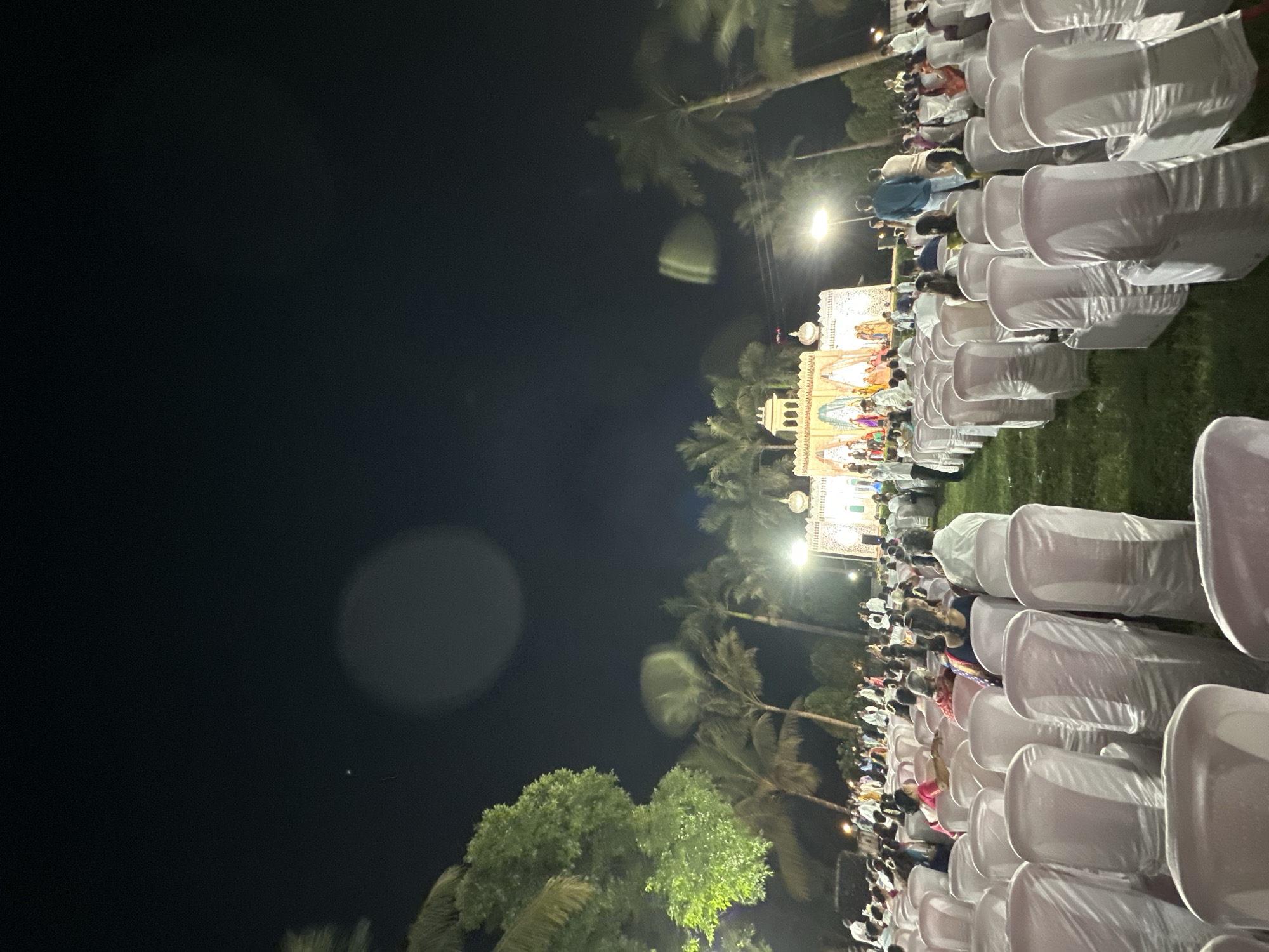 The image depicts an outdoor event at night, possibly in Shahapur, India, given the presence of palm trees and building style. Rows of white chair covers stretch into the distance where a crowd of people are gathered around what appears to be a brightly lit stage or building, possibly a temple or decorative structure. The attendees seem to be observing the stage or activity, but there is not enough detail to determine their exact actions. The night sky is dark, with some visible light interference. Green trees are situated at the lower left corner.