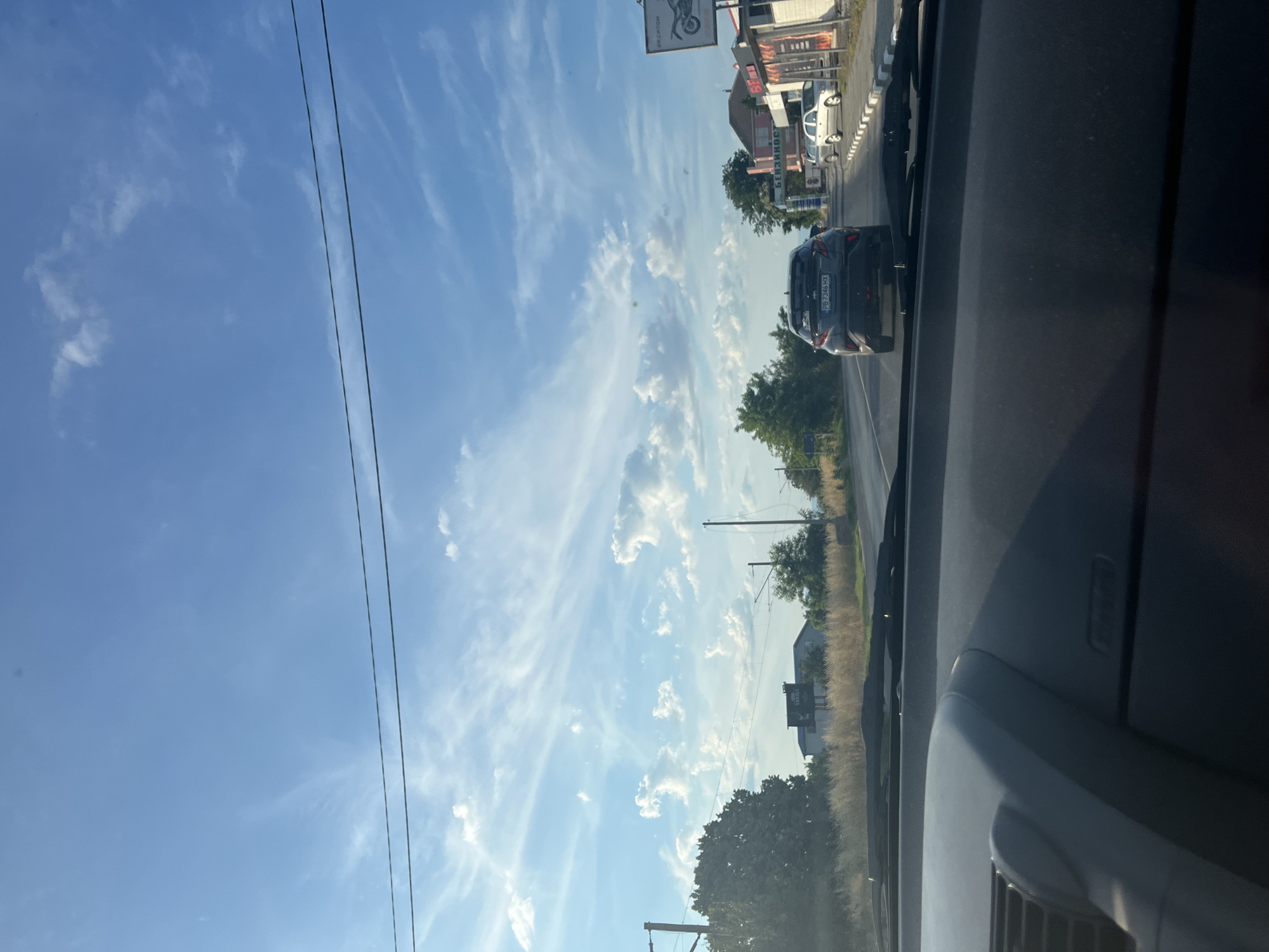 The image was taken from the interior of a vehicle traveling on a road in Trud, Bulgaria. The foreground shows the dashboard of the car, with the windshield extending upwards. Outside the vehicle, the road stretches ahead, with a dark-colored hatchback in front, displaying a license plate with the characters "PB 7346 HX". To the right of the road, there are several buildings, including a gas station with a sign reading "БЕНЗИНОСТАНЦИЯ" and another building with a prominent sign featuring a motorcycle. Further down the road, other vehicles are visible. The environment is characterized by a clear blue sky with scattered wispy clouds, and on either side of the road, there are trees, tall dry grass, and utility poles.